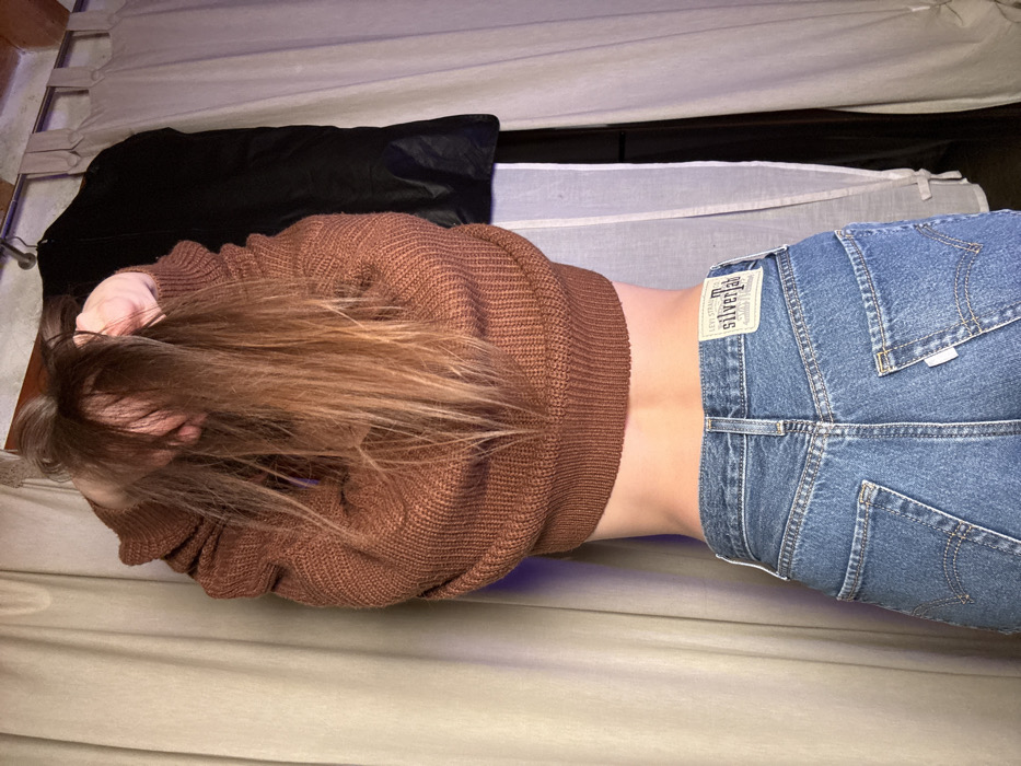 The image depicts an indoor scene in Monsummano Terme, Italy. A person is viewed from the back, from the waist up. The subject has long, light brown hair, which is held aloft by both hands at the crown of the head. They are wearing a rust-colored, ribbed knit sweater, which is lifted to expose the lower back. Blue denim Levi's SilverTab jeans are worn on the lower body, with a white and blue brand label visible on the rear waistline. The immediate background features light-colored curtains on both the left and right sides. A dark garment, consistent with a coat or suit bag, hangs from a metal rod positioned above the right-side curtain. Exposed rustic ceiling or wall material is visible in the upper background. The lighting is dim, with a subtle purple hue in the upper left area.