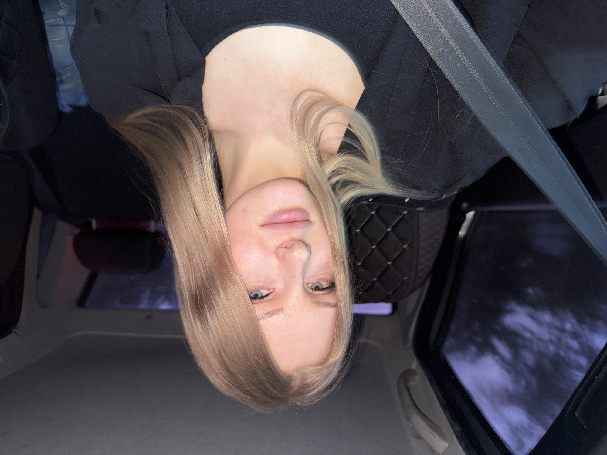 The image shows a fair-skinned blonde woman with light blue eyes in a car. She is wearing a black top and a gray seatbelt. The car's interior includes a black quilted seat cover. Through the window, there is a blurred, snowy, and likely urban environment, suggestive of the city of Perm, Russia. The image is rotated.