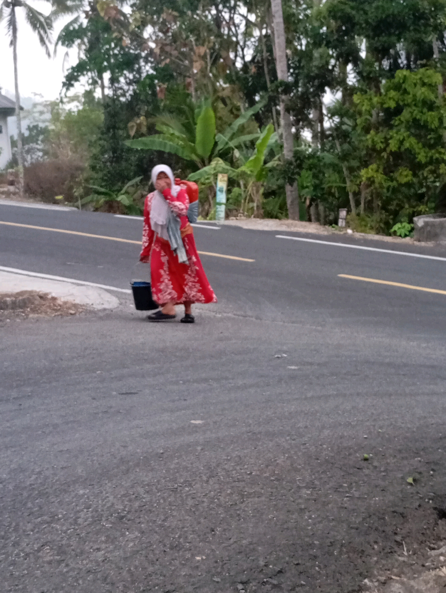 In Ketepung, Indonesia, a woman dressed in a red and white patterned dress and a light pink hijab walks on a paved road. She is carrying a red bag on her back and holding a black bucket with her left hand. The road is marked with white and yellow lines. The surrounding area is lush with trees and vegetation, and a building is partially visible to the left. The lighting suggests it is either early morning or late afternoon.