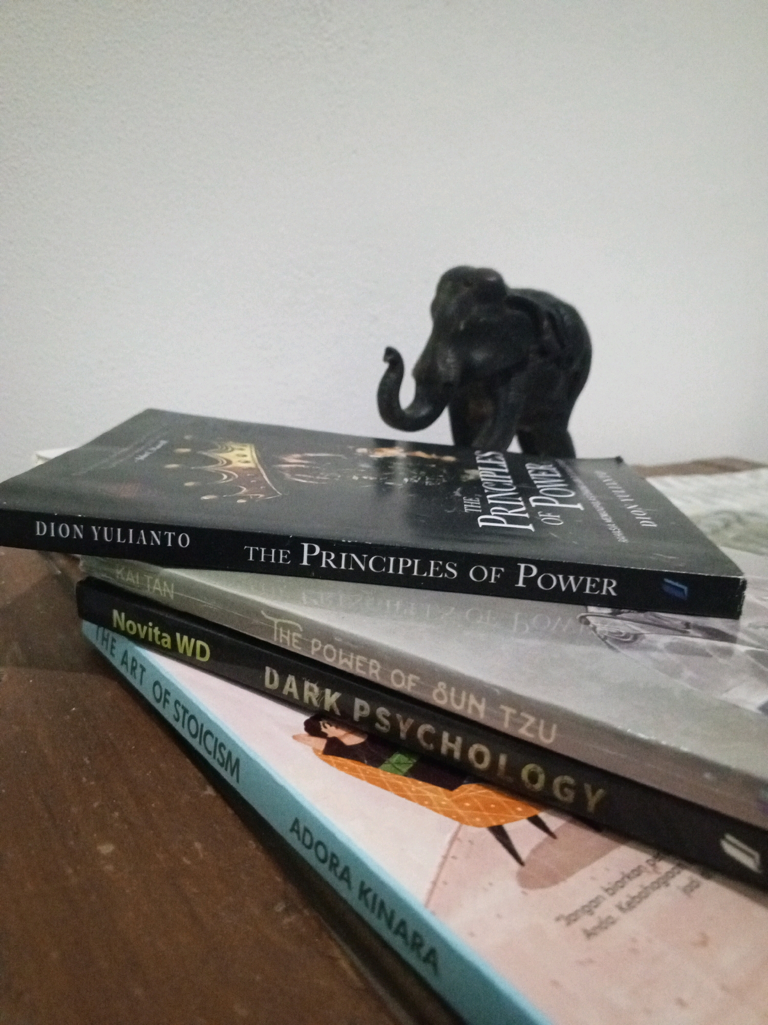 The image depicts a stack of books with a small dark elephant figurine on top. The books are arranged diagonally, with the top book being black and titled "THE PRINCIPLES OF POWER" by Dion Yulianto. Below it is a book with a lighter cover, partially visible, and then another black book titled "DARK PSYCHOLOGY" by Novita WD, also featuring "THE POWER OF SUN TZU" on its spine. The bottom book is light blue and titled "THE ART OF STOICISM" by Adora Kinara. The scene appears to be indoors, with a plain white background behind the books and a wooden surface beneath them. The lighting is soft, casting subtle shadows. No people are present in the image. The overall impression is a still life arrangement of intellectual material. There is no identifiable information in the image to place it in Denpasar, Indonesia.