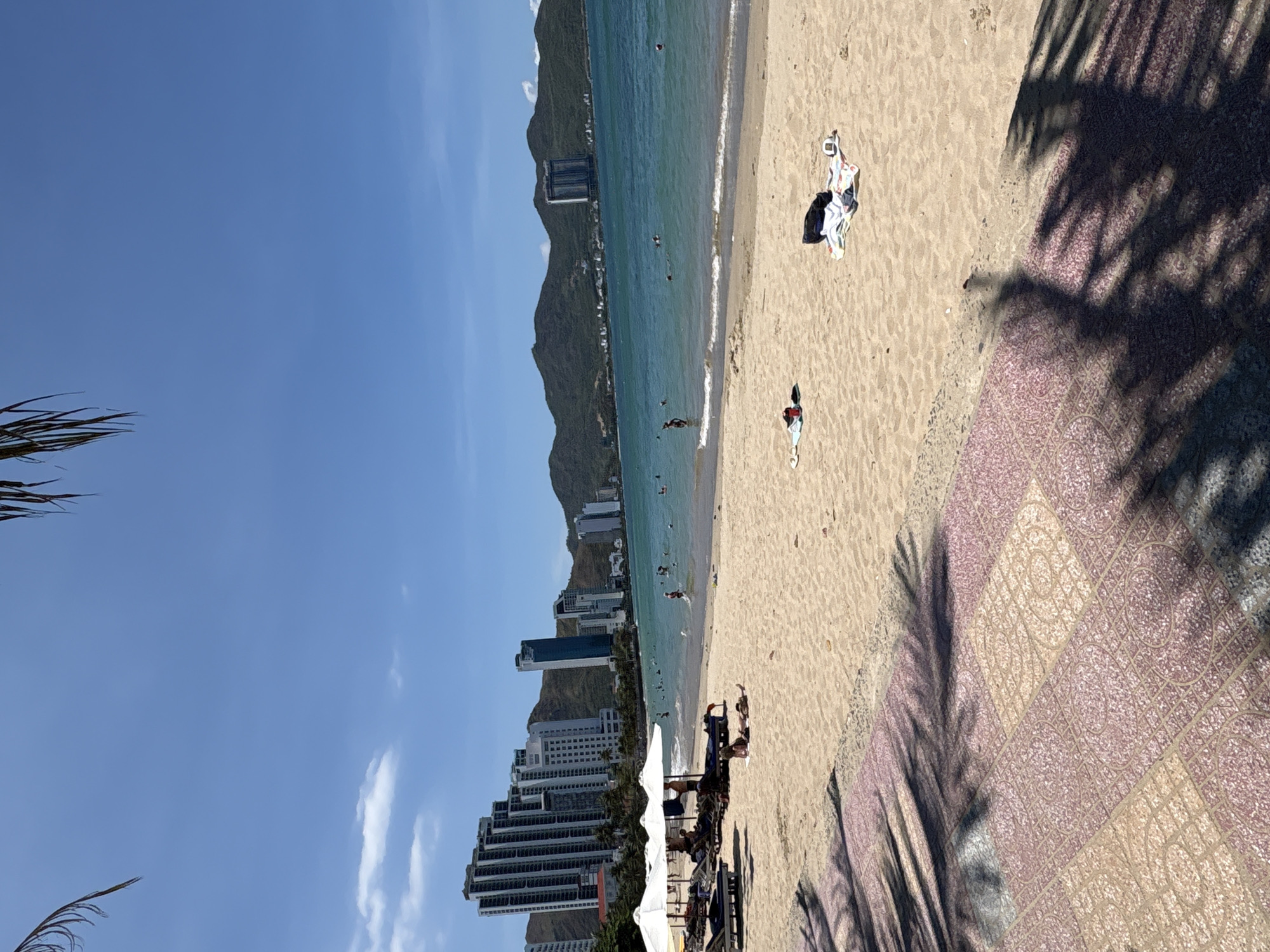 The image depicts a sunny day at a beach in Bac Nha Trang Ward, Vietnam. The scene includes a sandy beach with people relaxing, swimming, and sunbathing. In the background, a mountain range is partially obscured by high-rise buildings, some of which are hotels and apartments. In the foreground, there are shadows from palm trees cast over a tiled pathway adjacent to the beach. There are beach umbrellas and lounge chairs set up on the sand. The ocean appears calm with small waves. The sky is blue with a few scattered clouds. Some clothing and belongings are left on the beach.