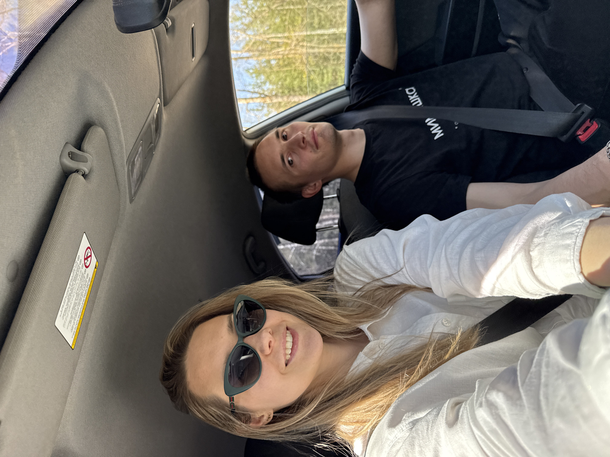 Two individuals are inside a vehicle. A woman with sunglasses and a white shirt is in the foreground. Behind her, a man with a black shirt and a seatbelt is visible. The vehicle is moving, as indicated by the trees visible through the window. The location may be Chaikovsky, Russia.