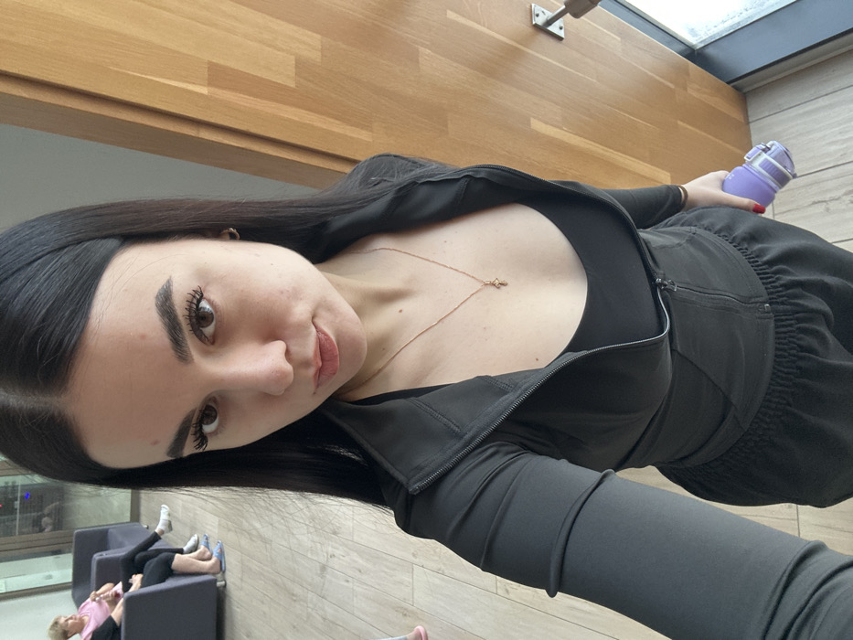 An indoor scene depicts a young woman in the foreground taking a selfie. She has long, dark, straight hair, dark eyes, and prominent eyebrows, looking directly at the camera. She wears a black long-sleeved athletic jacket, zipped partially open to reveal a black top underneath, and black athletic sweatpants. A thin gold-colored necklace with a small cross pendant is visible on her chest. Her right hand, partially visible at the bottom right, holds a purple and white water bottle.
In the background, to the left, two older women are seated in dark grey armchairs. One woman, partially visible, wears a pink top, light-colored pants, white socks, and light blue slippers. The other woman is further back and less distinct. The floor is covered with light-colored wooden laminate planks. A vertical wooden panel wall or door is visible on the right side of the frame. The environment suggests a public or commercial interior, possibly a waiting room or lounge in a fitness center, spa, or medical facility.
Based on the visual information presented, a specific location or identifiable landmark within the city of Saint Petersburg, Russia, cannot be determined.