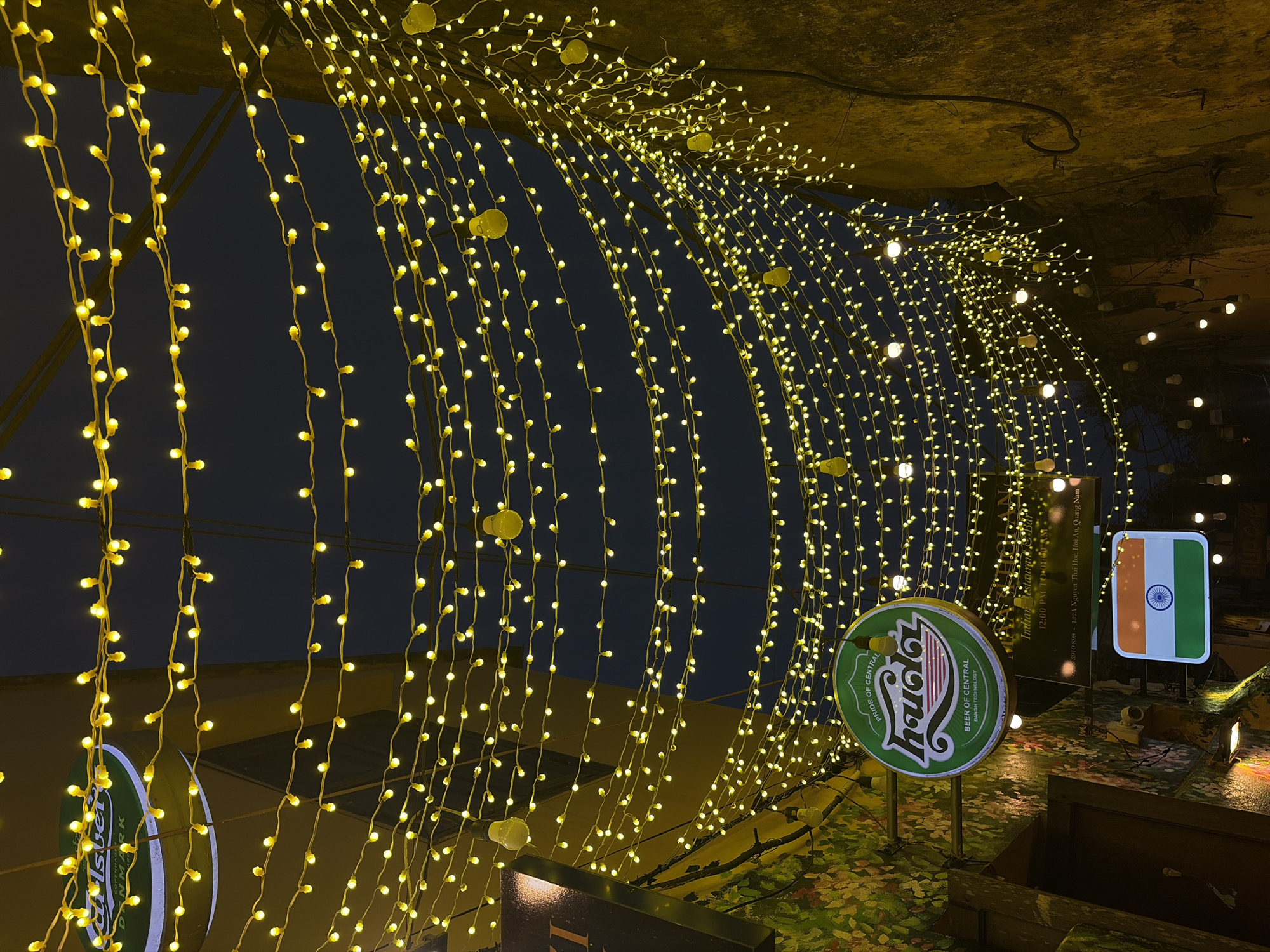The image presents a vibrant night scene, likely an outdoor pathway or entrance to a restaurant, illuminated by numerous warm yellow LED string lights. The lights are arranged in cascading curtains and arched patterns overhead, creating a festive and inviting tunnel-like canopy against a dark background.
Several prominent signs provide details about the location. On the left, a round green sign with a white logo and text is visible, reading "HUDA" and stating "PRIDE OF CENTRAL," "BEER OF CENTRAL," and "DANISH TECHNOLOGY." To its right, a rectangular sign prominently displays the flag of India, with its saffron, white, and green stripes and a blue Ashoka Chakra. Below the Indian flag, another rectangular sign, dark with white lettering, identifies the establishment as an "Indian Restaurant & Bar." This sign also includes practical information: operating hours from "12:00 PM TO 01:00 AM," a phone number "3910 899," and the address "132A Nguyen Thai Hoc, Hoi An, Quang Nam." A partially visible "Carlsberg" sign can also be seen.
The ground or a decorative raised platform in the foreground features an artificial turf-like surface with green and brown leafy patterns. The overhead structure supporting the lights appears dark and textured. The setting is clearly nighttime, emphasizing the glow of the artificial lights, and creates a cozy and welcoming atmosphere. The absence of people focuses attention on the elaborate light display and signage, pointing to an evening establishment in Hội An Ward, Vietnam.