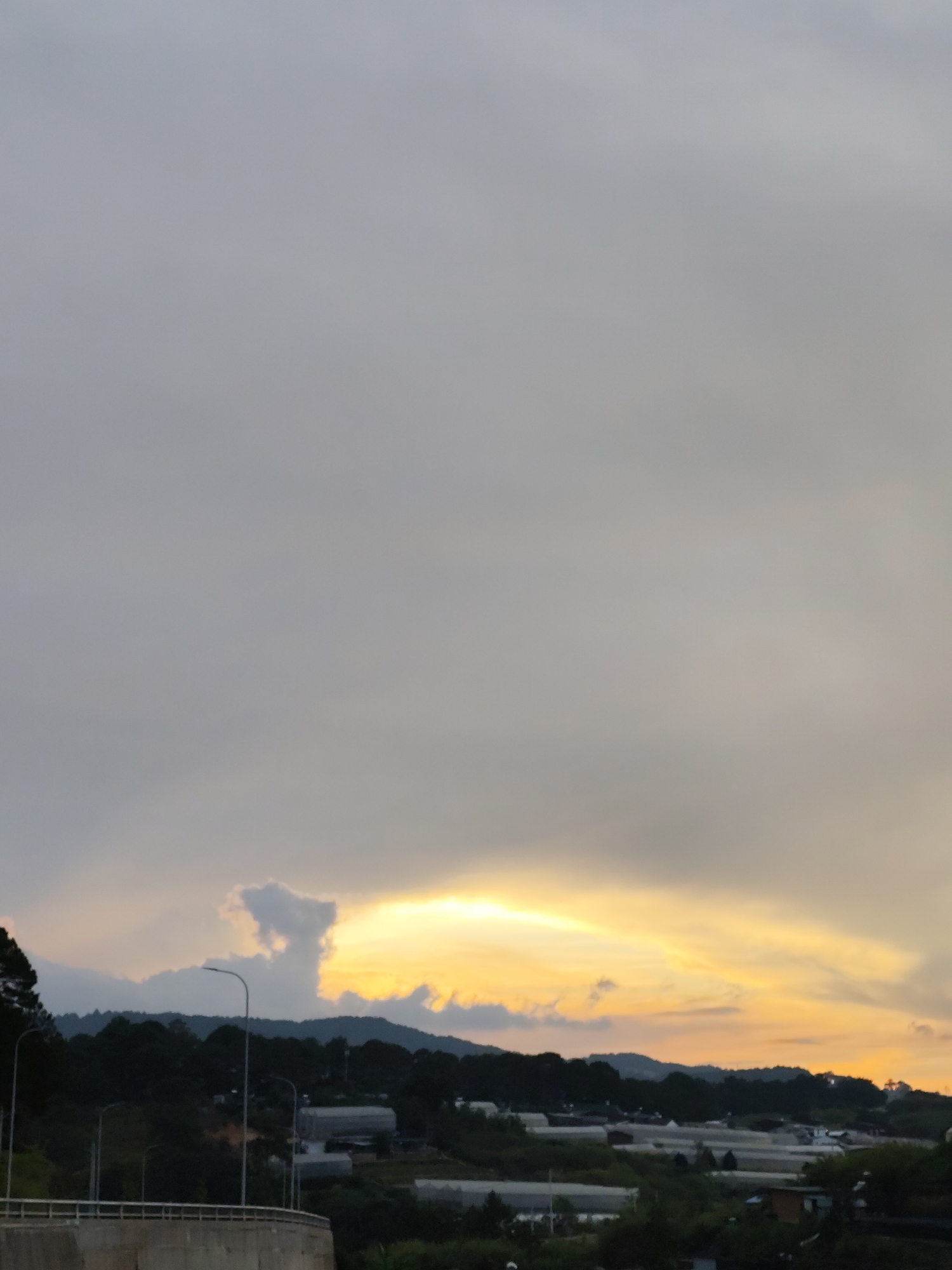 The photograph shows a landscape in Da Lat, Vietnam, with a cloudy sky dominating the upper portion of the frame. The lower section features a silhouette of a forested hill and what appears to be a residential area with greenhouses or similar structures. A concrete barrier or wall is visible in the foreground. Streetlights are positioned along a road or path near the trees. The sky exhibits a gradient of colors, with areas of yellow and orange near the horizon, suggesting either sunrise or sunset.