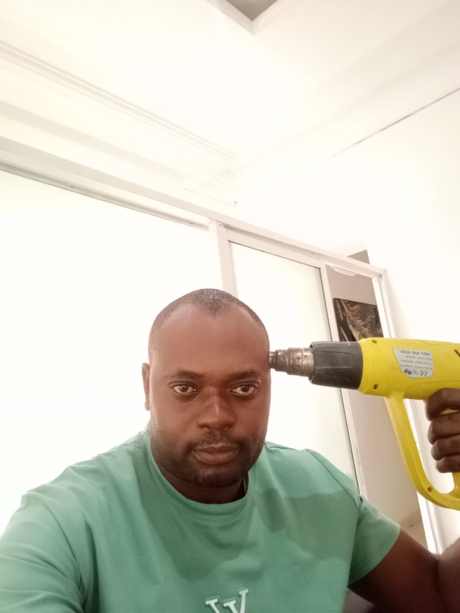 A man is posing for a selfie indoors. He is holding a yellow heat gun to his head. He is wearing a green t-shirt. The background includes a white wall with a window frame. The image was presumably taken in Gwarinpa, Nigeria.
