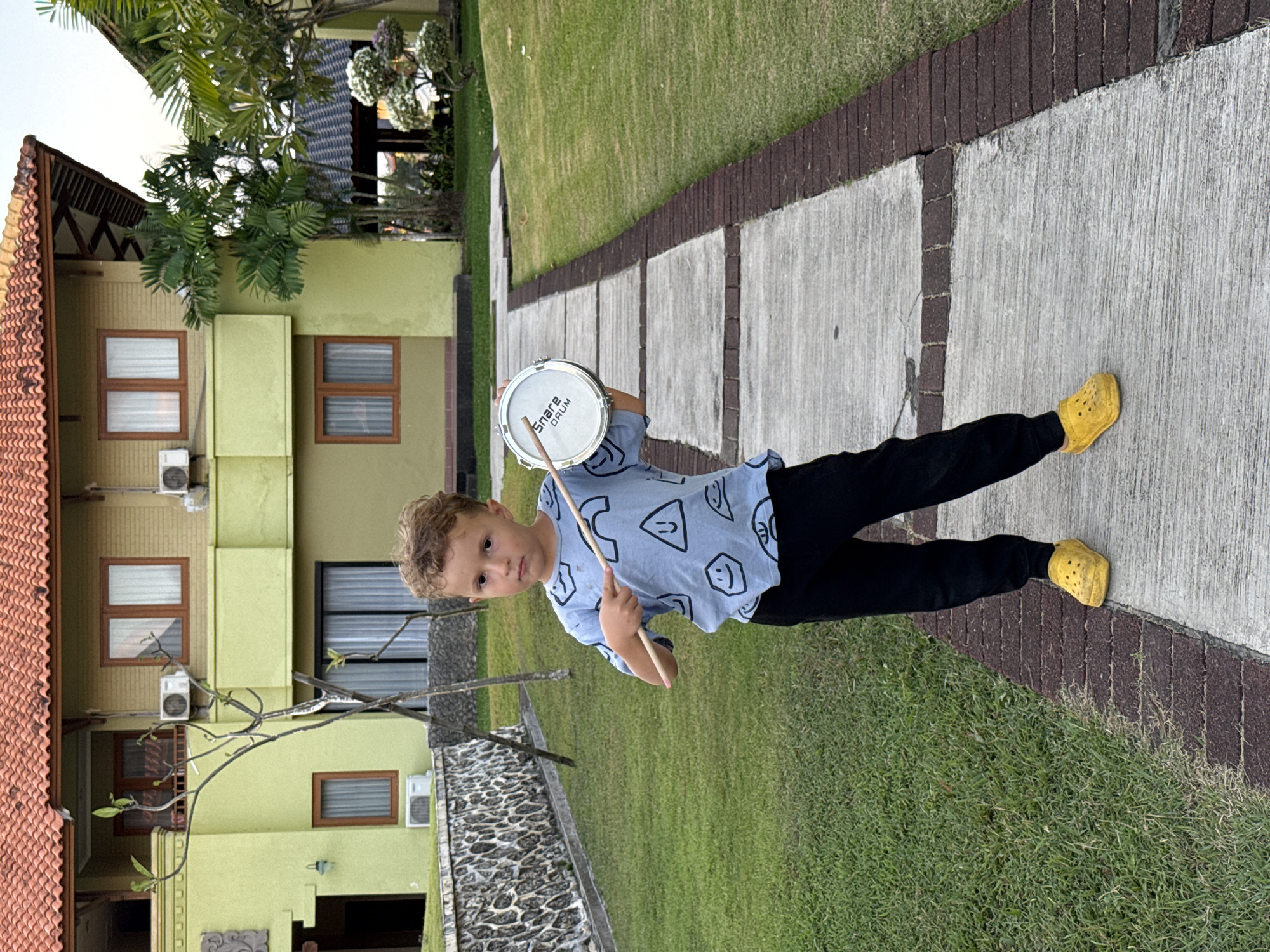 A young boy stands outdoors, holding a snare drum and a drumstick, looking directly at the camera. He has light brown curly hair and is wearing a light blue t-shirt with black, stylized face graphics, black pants, and bright yellow Crocs. The boy holds a small, white snare drum in his left hand, with a wooden drumstick in his right, poised over the drum. The words "Snare Drum" are clearly visible on the drum head.
The setting features a light green two-story building in the background, distinguished by a reddish-brown tiled roof and numerous rectangular windows with brown frames. Several air conditioning units are mounted on the building's exterior. The boy is standing on a light grey concrete path, which is bordered by dark brown bricks and flanked by lush green grass on either side. Palm trees and other tropical foliage are visible, suggesting a warm, tropical climate. The image appears to be taken during the daytime, possibly late afternoon, with clear lighting. The scene is consistent with an outdoor area, possibly in Kuta Selatan, Indonesia.