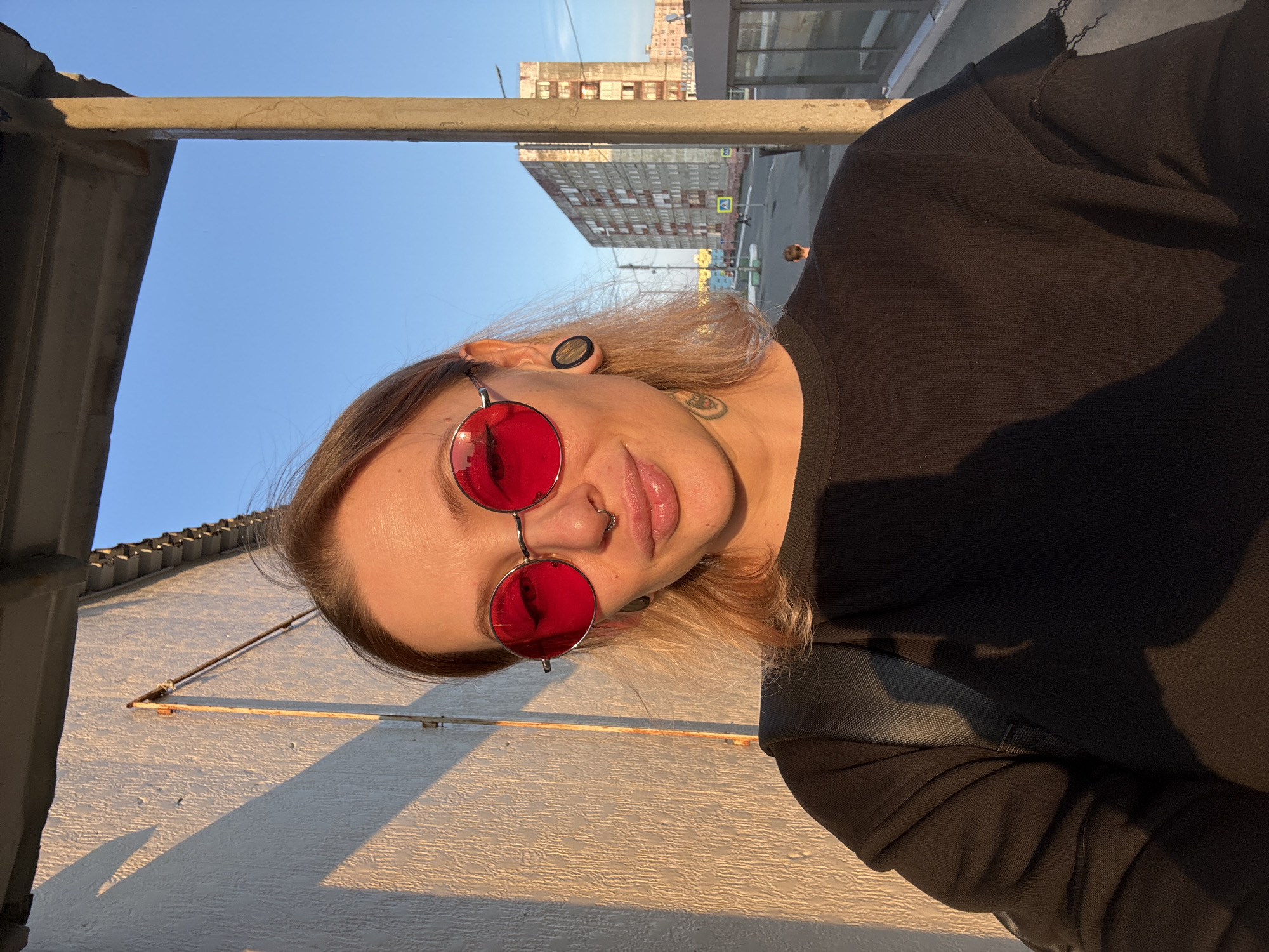 The media file is a portrait of a person wearing round red-tinted sunglasses and a black long-sleeved shirt. The person has blonde hair and visible ear gauges. A septum piercing is also visible. The image appears to be taken outdoors during daylight, as indicated by the bright sunlight casting shadows. The background shows a clear blue sky, some buildings, and a street. The person is looking directly at the camera with a neutral expression. The overall tone of the image is casual and informal.