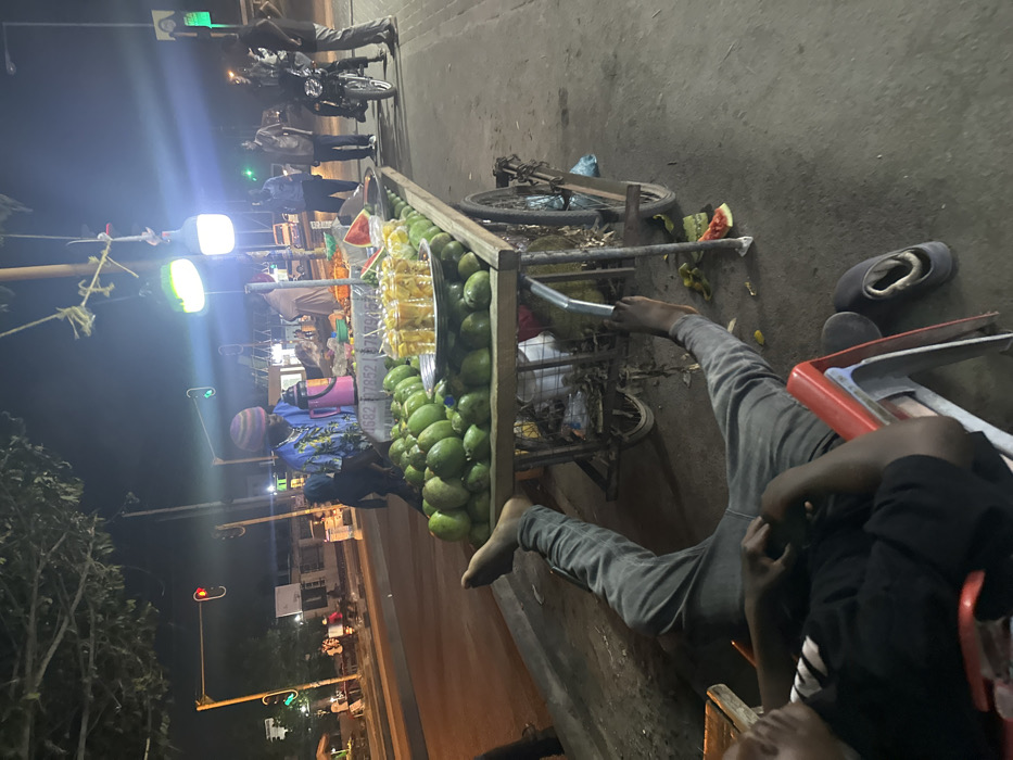 A street scene at night shows a fruit cart laden with green mangoes, watermelon, and cut fruit. People are milling around the cart. A man reclines in a chair, his bare feet near the back of the cart. A bicycle leans against the cart. Streetlights illuminate the scene. A street stretches into the distance, with buildings and trees visible. There's a traffic light and some parked motorcycles.