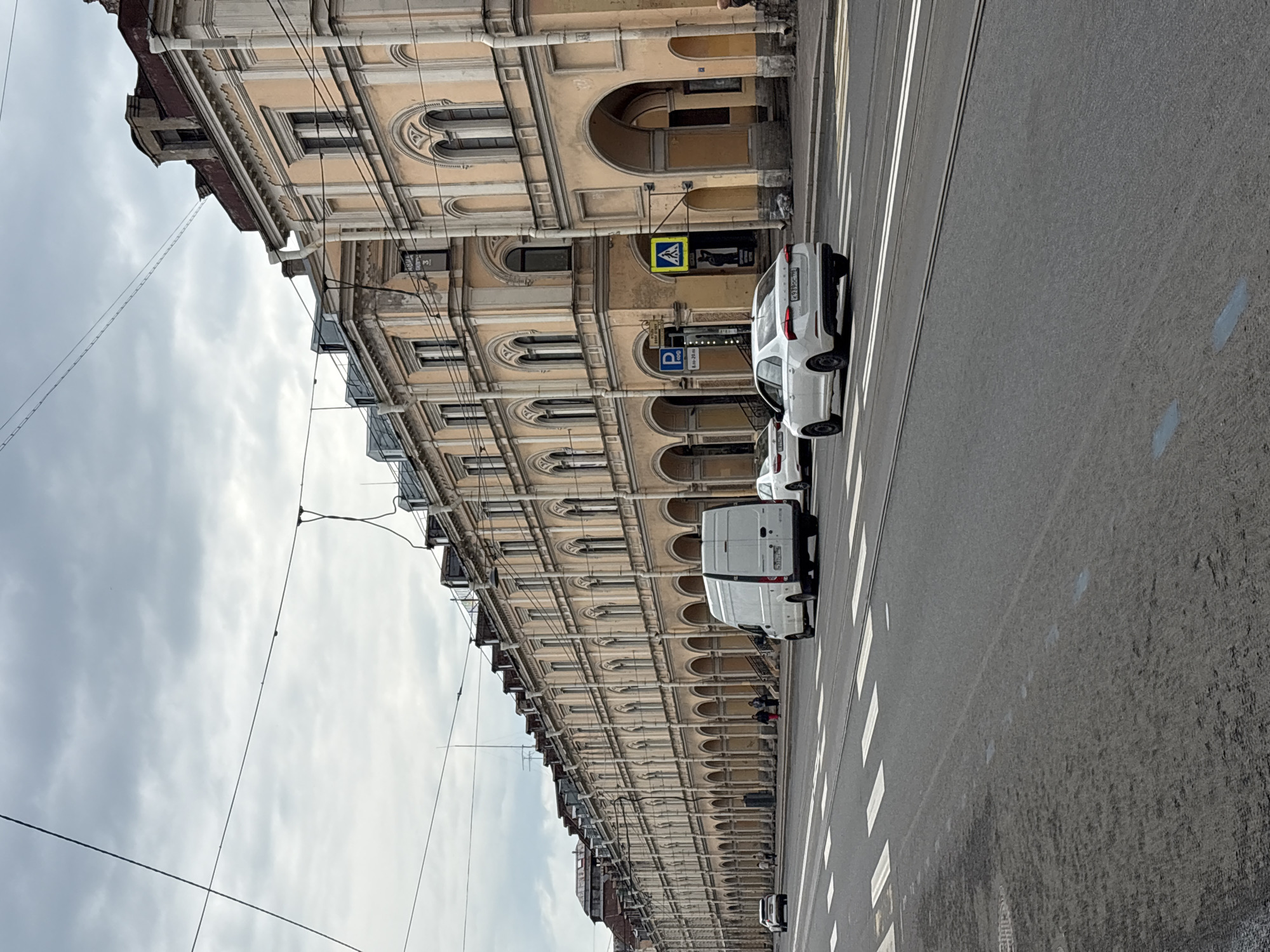 The image depicts a street scene in Saint Petersburg, Russia. The architecture is characterized by a long, ornate, yellow-toned building with arched windows and colonnades running along its facade. This building is on the left side of the image. The street has a pedestrian crossing indicated by white stripes. There are several vehicles parked along the roadside, including a white van and at least one white car. There is street signage visible, including a pedestrian crossing sign and a parking sign. Overhead, there are numerous power lines crisscrossing. The sky is overcast with grey clouds.