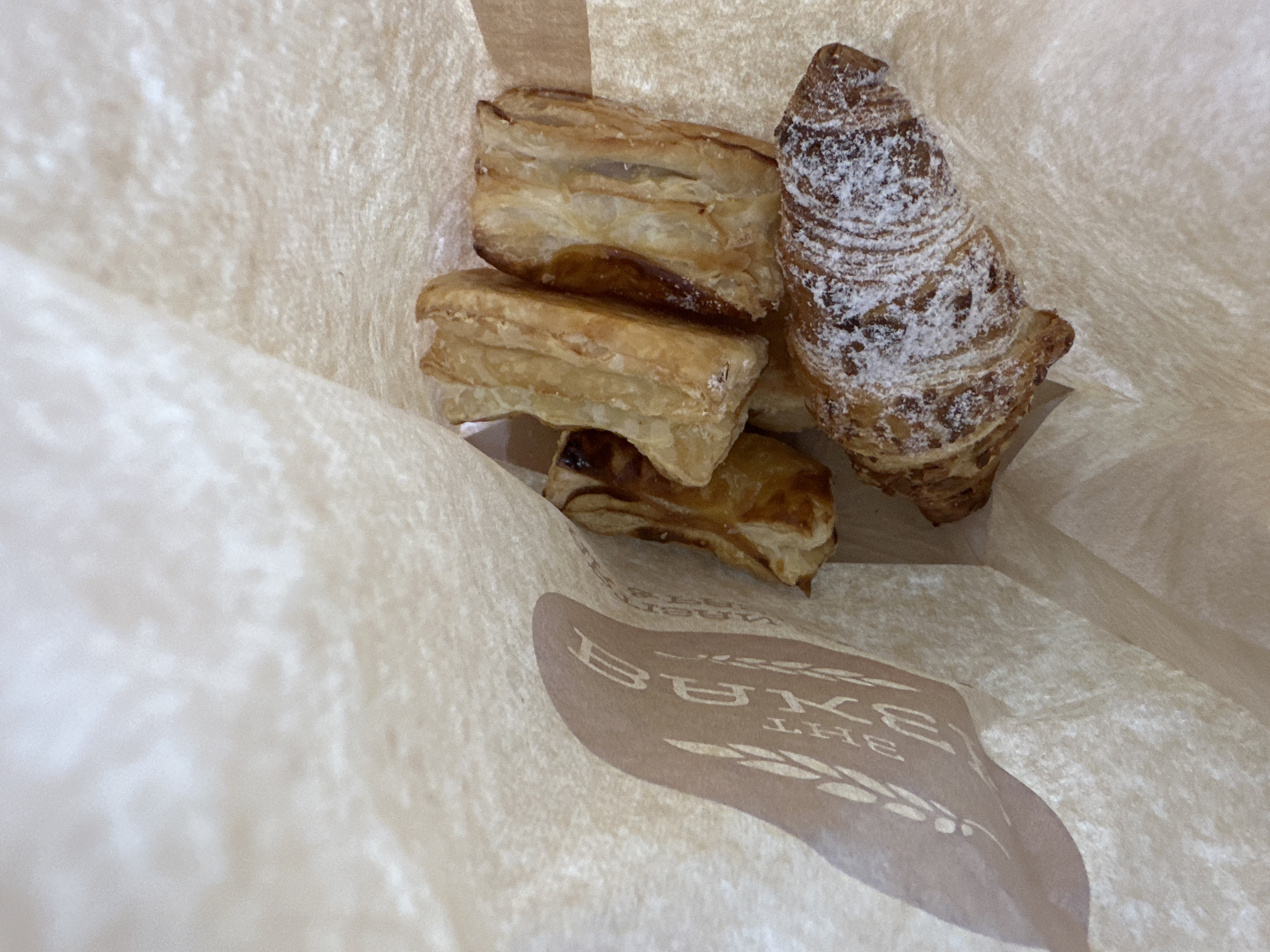 A paper bag is open and facing upwards. Inside the bag, there are five baked goods. One is a croissant dusted with powdered sugar. The other four are puff pastry pastries, appearing to be filled with something. The bag has a logo printed on the inside flap with wheat stalks and text that reads "BAKE THE...". The location is the bakery in Tambon Ko Pha Ngan, Thailand.