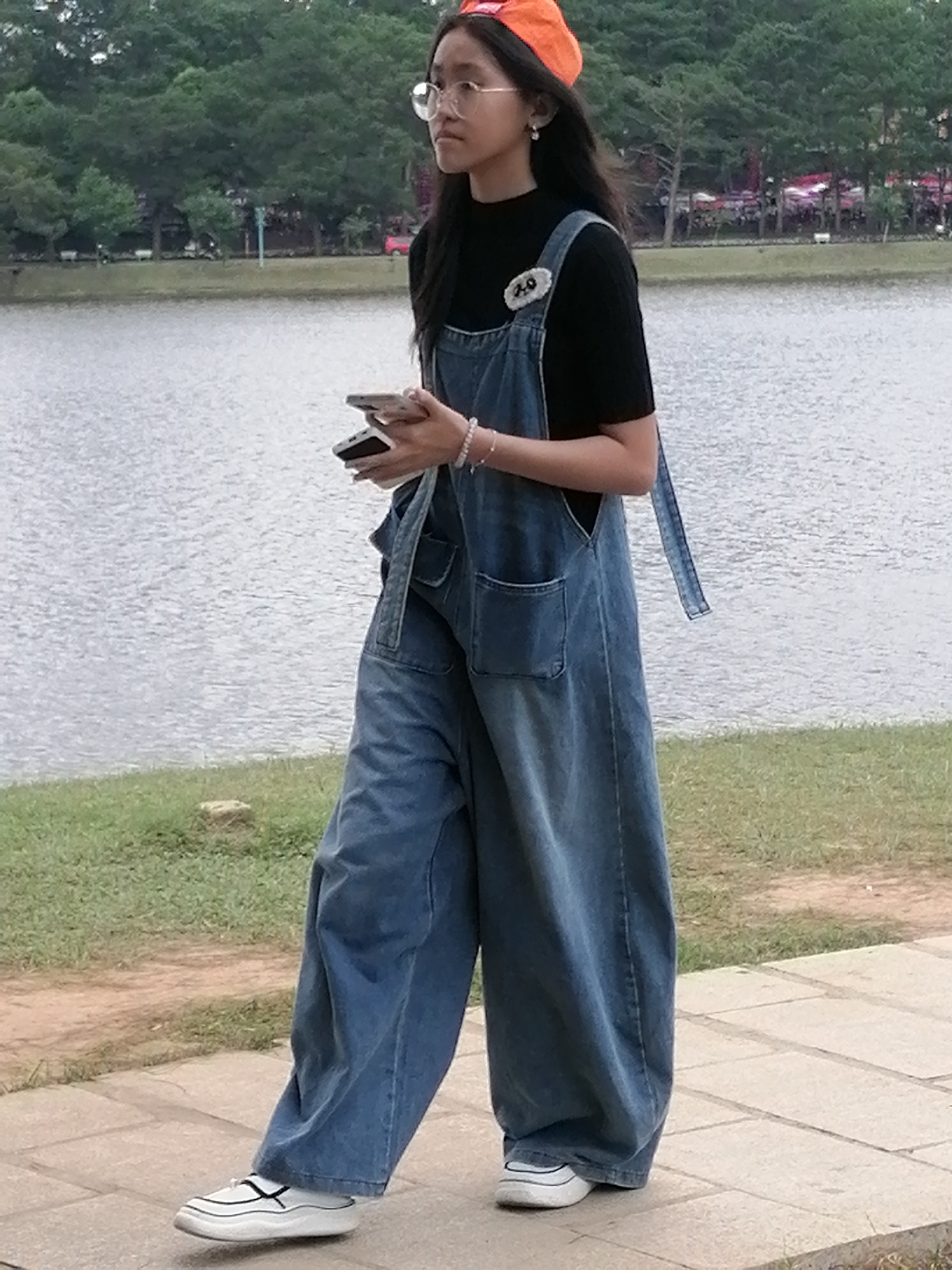 A young Asian woman stands on a paved walkway near a body of water. She is wearing a black long-sleeved shirt, blue denim overalls, an orange cap, and glasses. She is holding two cell phones in her hands and appears to be looking to her right. The background features a lake, trees, and some distant structures. The scene is likely in Da Lat, Vietnam, a city known for its natural beauty and cooler climate, often featuring lakes and pine forests.