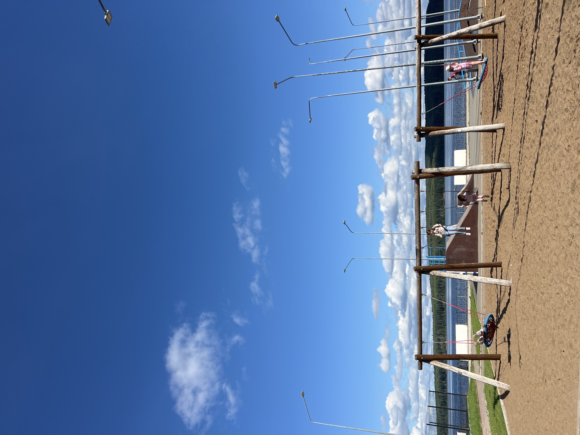 The image captures a sunny day at a playground located on the bank of the Kama River in Chaikovsky, Russia. The foreground is dominated by a sandy playground with several wooden swing sets. In the center, a round, net-like swing holds one child. To the right, two more children are on individual swings; one is wearing a pink outfit and a hat, and the other is wearing a pink outfit. A person in a white top and jeans is standing near a ramp in the background.
The middle ground features the calm blue waters of the Kama River, bordered by a dense green forest on the opposite bank. A fence runs along the river's edge.
The background is a vast, bright blue sky with scattered white clouds. Numerous tall, silver lampposts are positioned along the horizon, suggesting the area is equipped for evening use. The overall scene depicts a recreational area designed for outdoor activities, set against a natural landscape.