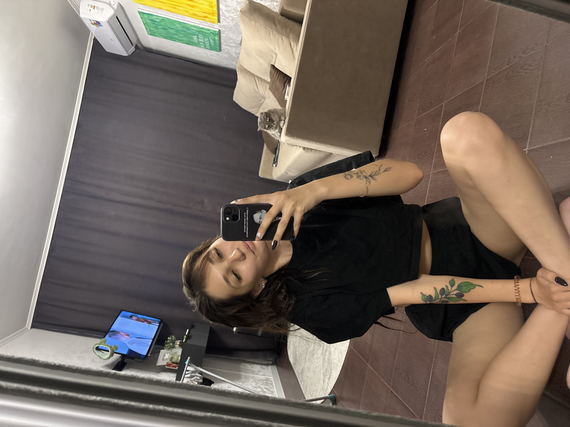 A young woman is seated on the floor in a room, taking a mirror selfie. She is wearing a black crop top and black shorts. She has long, dark hair and is wearing black nail polish. She has a tattoo of a branch with leaves and berries on her left arm and another tattoo on her right arm. A cat is visible on a beige sofa in the background. There is a television mounted on the wall, an air conditioning unit, and two framed pieces of art on the wall. The room has wooden flooring and dark grey curtains. The image appears to have been taken in an apartment in Novokuznetsk, Russia.