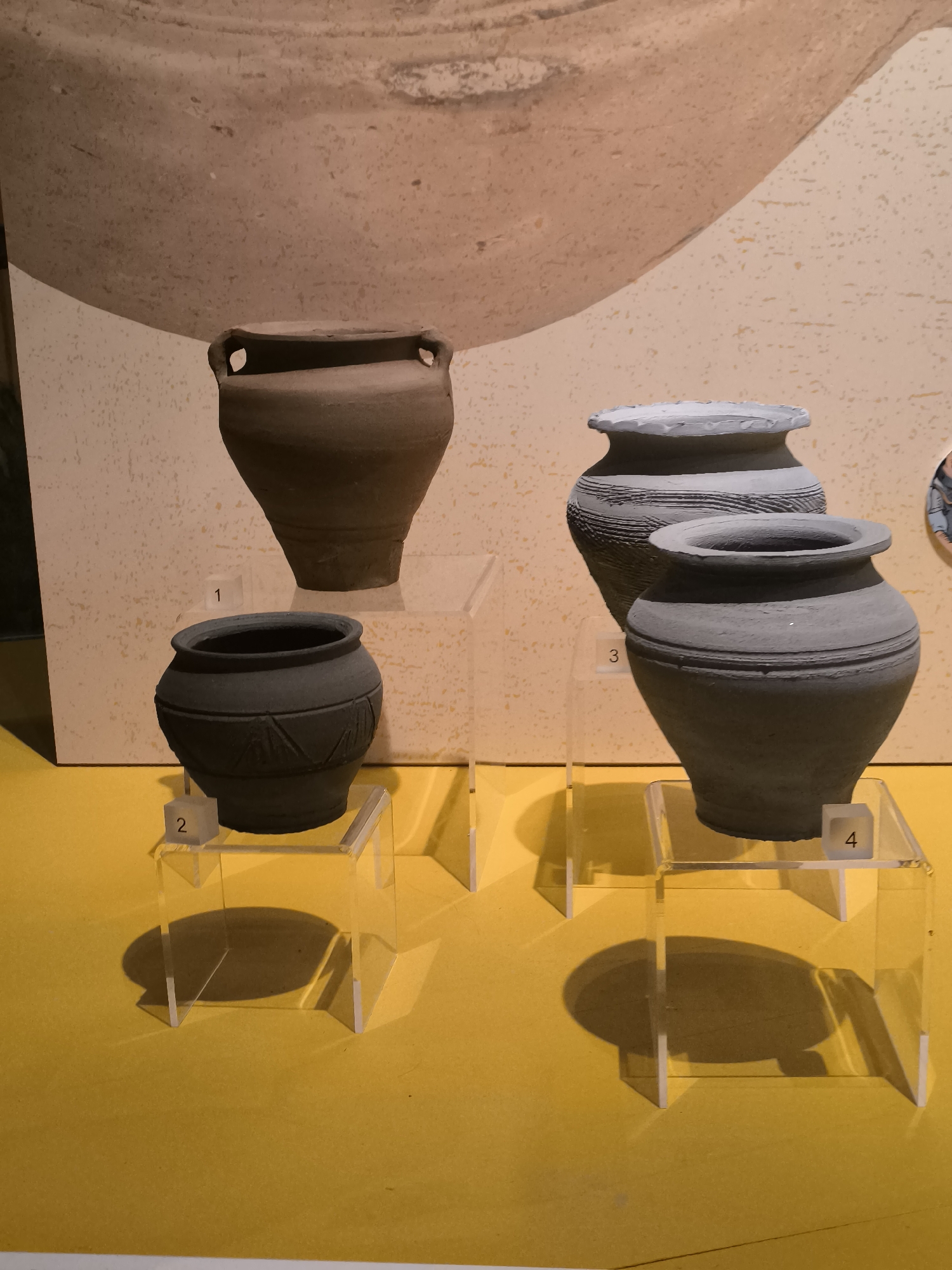 The image shows four ancient pottery vessels displayed on clear acrylic stands, numbered 1 to 4, against a backdrop depicting a larger pottery vessel. The vessels are of varying shapes and sizes, with numbers 2 and 4 being darker in color than numbers 1 and 3. The background is a beige-toned wall. The vessels are illuminated by overhead lighting, creating shadows beneath the stands. The floor or surface beneath the display is bright yellow. There is no visible evidence to confirm that it is at Castricum, Netherlands.