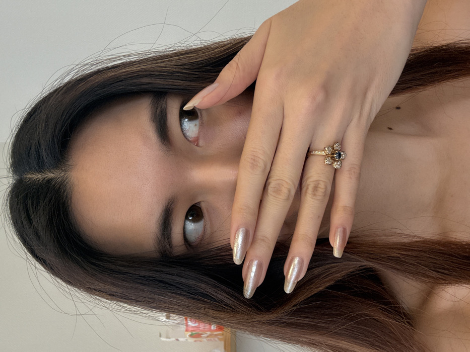 The image captures a close-up, chest-up view of a female individual located in an indoor setting within Arcueil, France. The subject, of East Asian descent, possesses long, dark brown hair with lighter undertones, parted centrally. Her brown eyes are visibly directed to the viewer's right. Her right hand is positioned horizontally across her lower face, covering her mouth and nose. The fingernails on her right hand are long, almond-shaped, and coated with a shimmering, iridescent light peach-gold polish. A gold-toned ring, featuring a central dark stone surrounded by smaller clear stones arranged in a floral pattern, is worn on the ring finger of her right hand. The subject's bare left shoulder is partially visible. The background consists primarily of a plain white wall, with indistinct, out-of-focus objects visible on the far left edge.