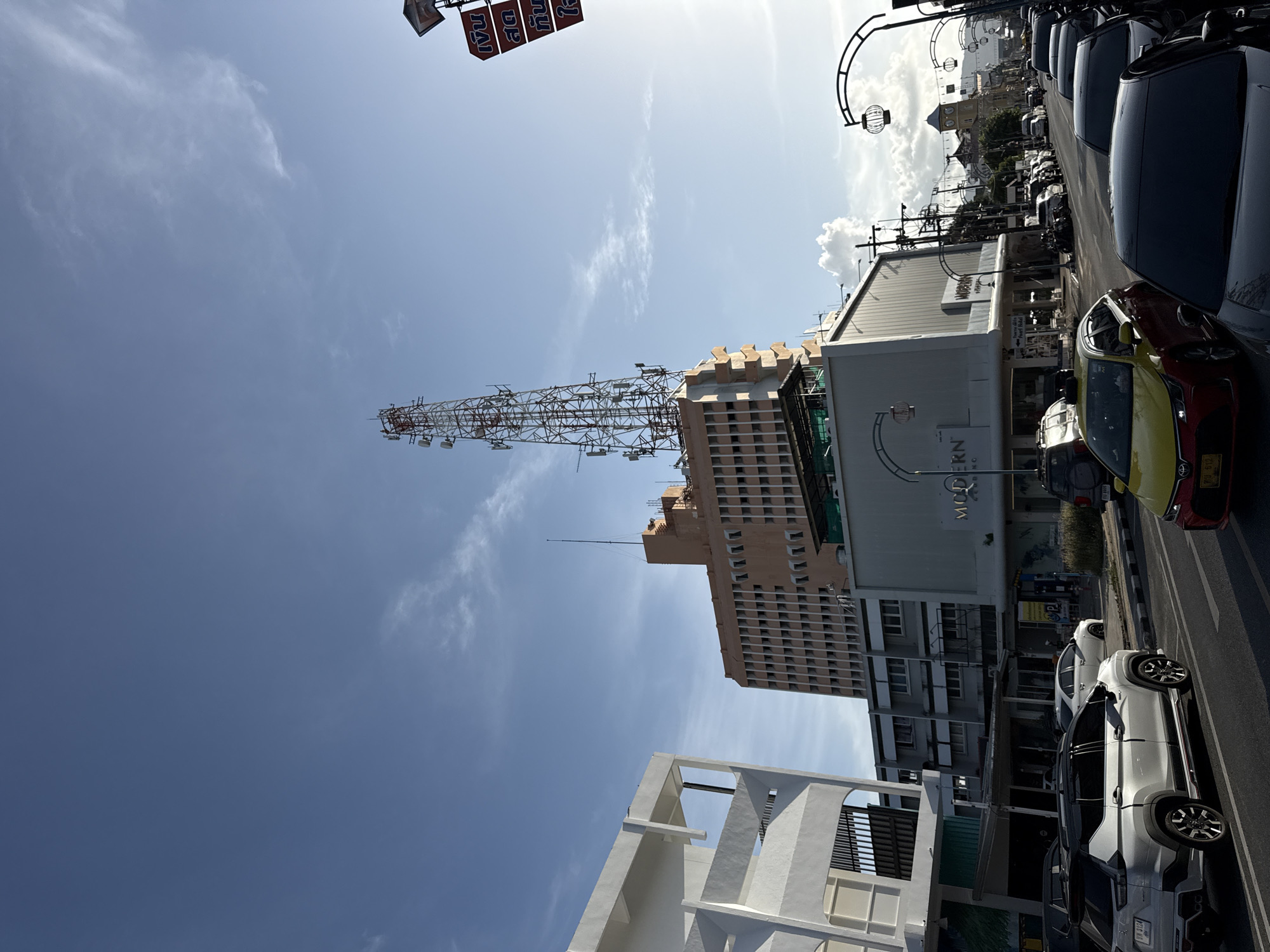 This image captures a street scene in Phuket City Municipality, Thailand, under a clear blue sky with scattered clouds. The foreground shows a road with several cars, including a silver SUV and a yellow and red taxi. The background features a row of buildings, one of which is a tall, peach-colored structure topped with a prominent telecommunications tower. Another building in the middle ground is a modern, white structure with the word "MODERN" visible on its facade. The street is lined with ornate streetlights, suggesting a developed urban area. The overall impression is one of a bustling city with a mix of commercial and residential buildings, and a significant telecommunications infrastructure.