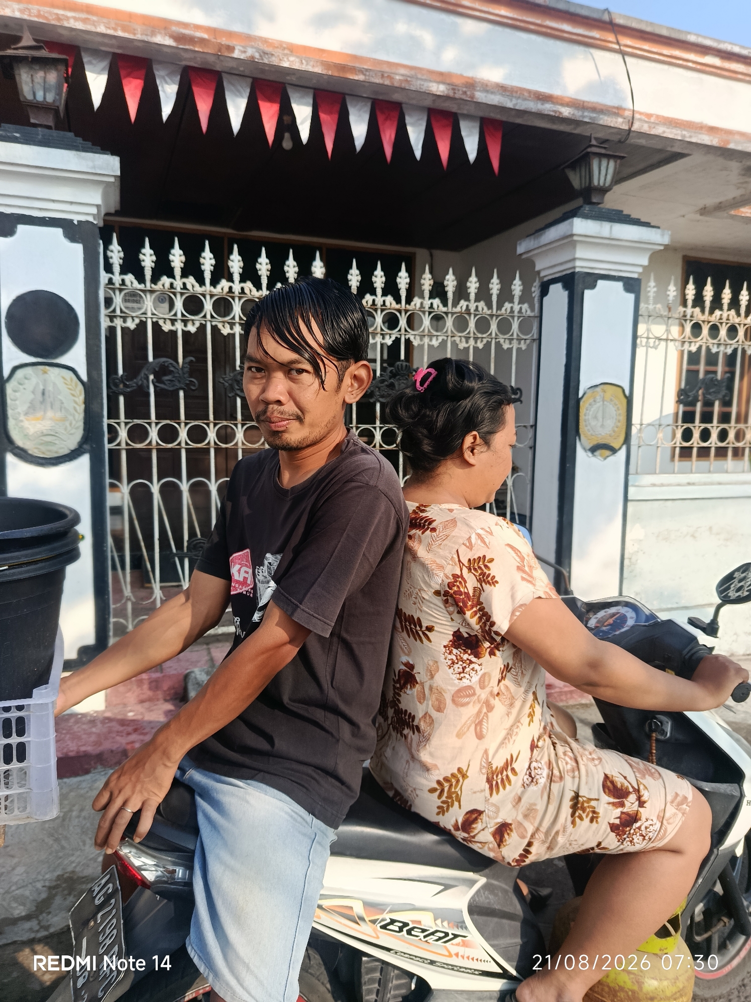 The image depicts a man and a woman sitting on a motorcycle in front of a house. The man, facing the camera, has short, wet dark hair and is wearing a black t-shirt and light blue denim shorts. He has a mustache and a short beard. The woman is sitting on the motorcycle behind him, facing away from the camera, with her dark hair pulled up and a pink clip. She is wearing a light-colored dress with a floral pattern. The motorcycle appears to be a scooter.
In the background, a white house with black and white trim is visible. Red and white pennant flags are strung along the roofline. A metal fence with decorative elements stands in front of the house, and there are potted plants or buckets on the left side of the image. The ground appears to be paved with concrete and some red-bricked steps. The scene is illuminated by sunlight, suggesting it is daytime. The timestamp "21/08/2026 07:30" and "REDMI Note 14" are visible in the lower right and left corners respectively, suggesting this is a photograph taken with a Redmi Note 14 phone on August 21, 2026, at 7:30 AM. The image is likely taken in Panggungrejo, Indonesia, as indicated by the prompt.