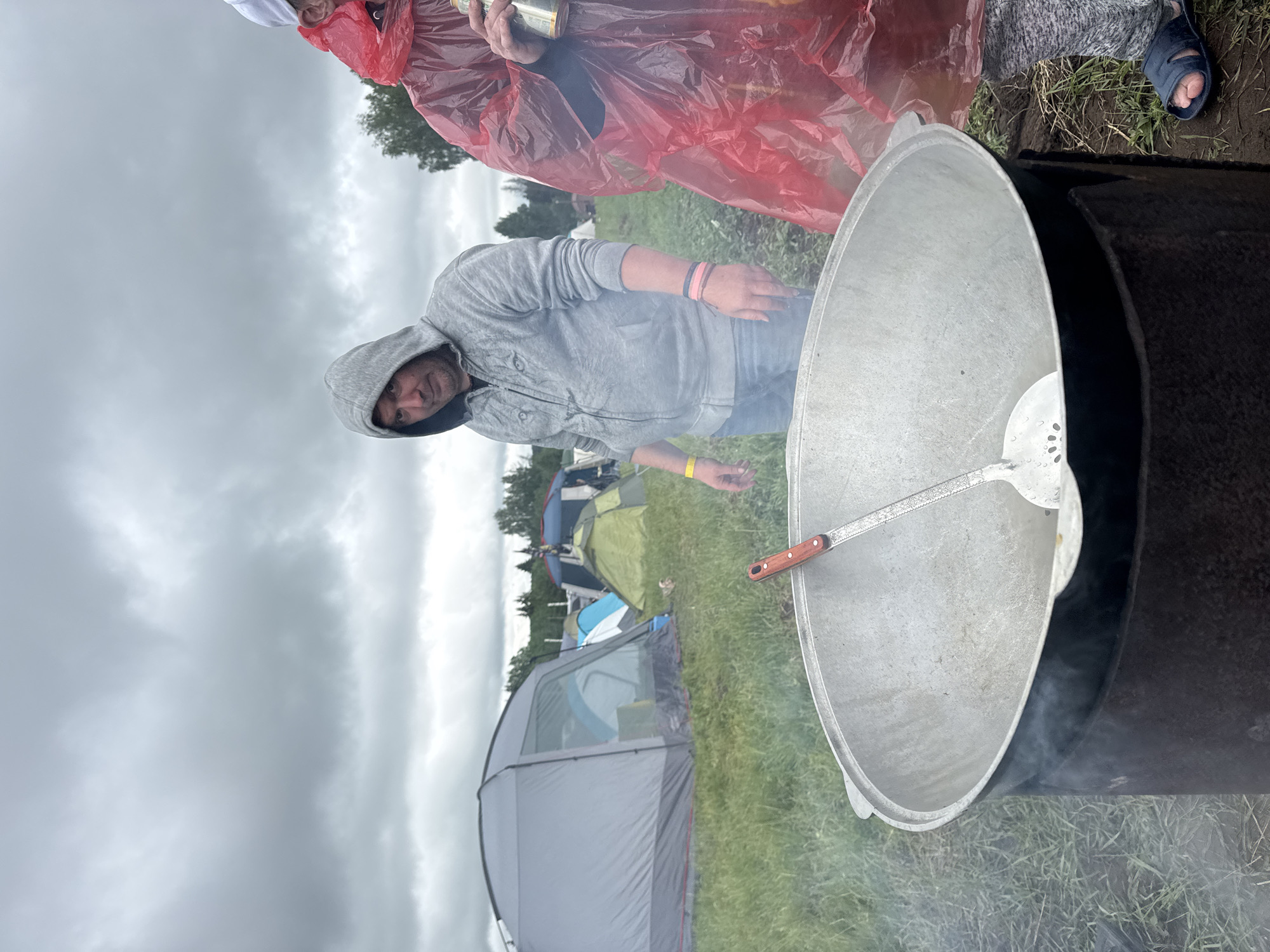 The image captures an outdoor scene under a heavily overcast, grey sky, presumably in Большая Кивара, Russia. In the foreground, a large, silver-colored metal cauldron (kazan) rests on a dark, cylindrical metal stove, from which white smoke emanates. A large metal slotted spoon with a wooden handle is placed diagonally inside the cauldron.
To the left of the cooking setup, a man wearing a grey hooded zip-up sweatshirt, blue jeans, a yellow wristband on his left arm, and multiple colored wristbands on his right arm, stands with his hands by his sides. He has short, dark hair and a beard/stubble, and is looking towards the viewer with a neutral expression.
To the right of the cauldron, a person is partially visible, wearing a bright red translucent plastic rain poncho with the hood up, a white baseball cap beneath the hood, grey sweatpants, and dark blue slip-on sandals. They are holding a silver beverage can in their right hand.
In the midground, several tents are pitched on a grassy field. A prominent grey dome tent is visible on the left, with its entrance slightly open. Behind it, a blue and white tent can be seen, and further to the right, a green dome tent and another blue tent are visible among scattered trees in the distance. The ground is a mix of green grass and patches of bare earth.