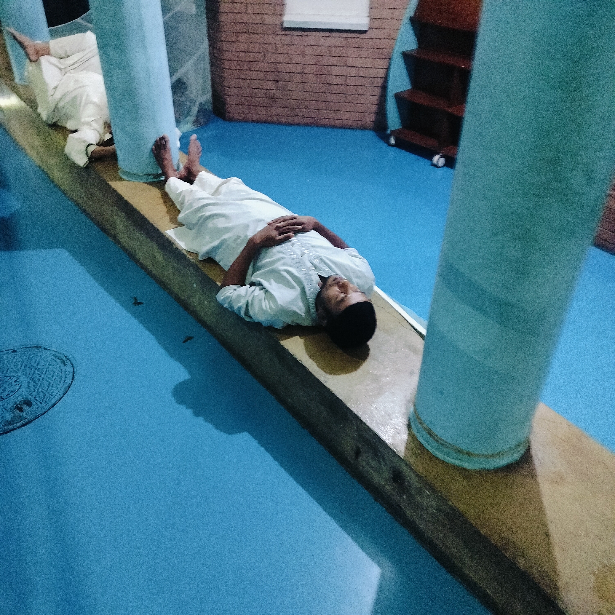 In Dhaka, Bangladesh, the interior space has a blue floor. A man, wearing a white garment and a black cap, is lying on a wooden beam between two cylindrical blue pillars. Another person in white is partially visible in the background. Behind them, there's a brick wall, a window, and a small shelving unit. A manhole cover is visible on the floor.