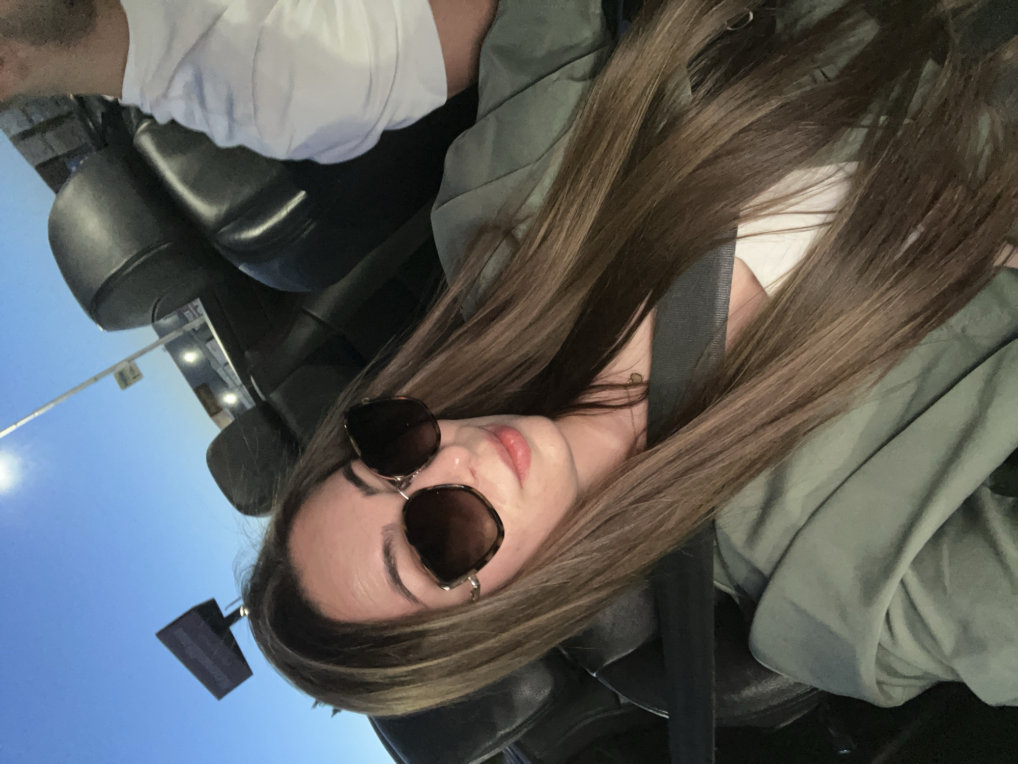 The image depicts a woman with long, brown hair wearing sunglasses and a green jacket, sitting in the passenger seat of a convertible. She is looking towards the camera. In the background, the sky is a clear blue, suggesting it is daytime or early evening. The car's black leather seats are visible, as is a portion of a white t-shirt worn by another person in the car. A seatbelt is visible across the woman's chest. The overall setting appears to be an urban environment, possibly a street or parking lot, with a lamppost and a billboard visible in the distance. The scene does not contain any specific elements that would allow for definitive identification of the location as Plovdiv, Bulgaria.