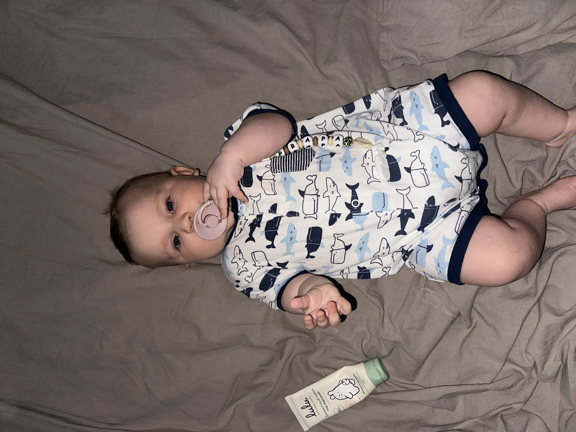 A baby wearing a white and blue shark-patterned romper is lying on a grey bedsheet. The baby has a pink pacifier in their mouth and a wooden bead pacifier clip attached to their romper. A white tube of "Lulu Natural Care" baby cream with a green cap is located near the baby's feet. The location is in Chaikovsky, Russia.
