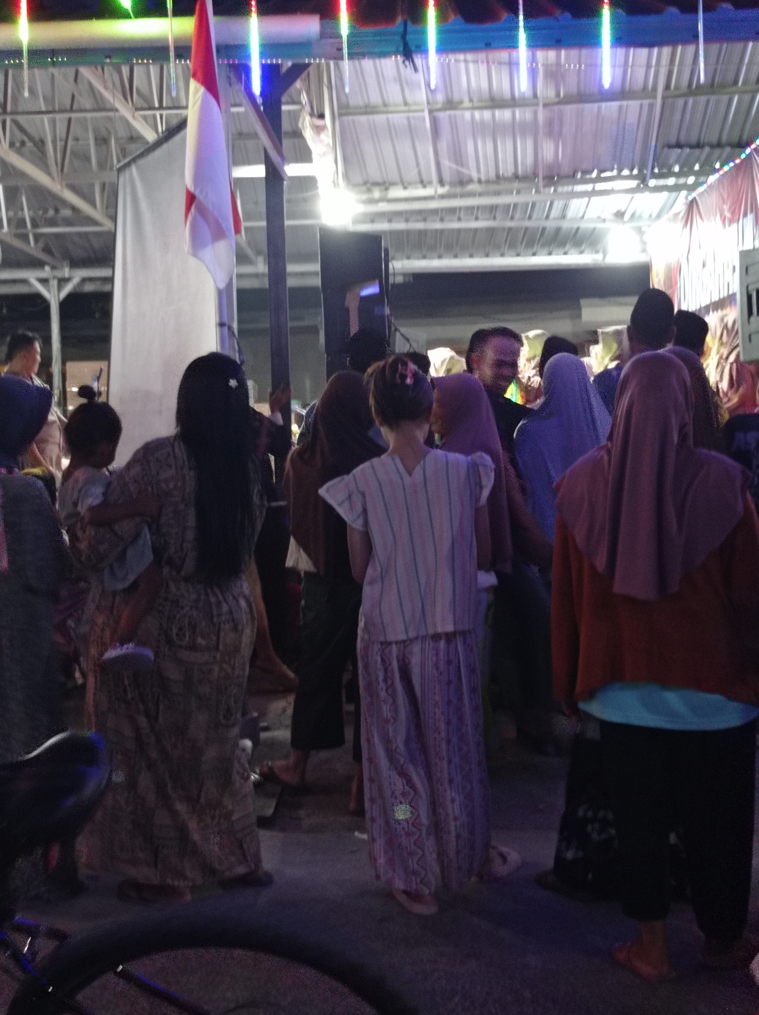 In Plumbungan, Indonesia, at night, a group of people gather under a metal-roofed structure adorned with colorful lights, suggestive of a public event or celebration. A flag with red and white horizontal stripes, likely the Indonesian flag, hangs prominently, adding a patriotic element to the scene. In the foreground, a woman is holding a child, both dressed in traditional attire. Other individuals in the crowd are also seen wearing headscarves and long garments. The lighting appears to be artificial, casting shadows and illuminating the event space. The ground is paved, indicating an outdoor or semi-outdoor urban setting. The atmosphere seems festive and communal, with people interacting and observing the proceedings.