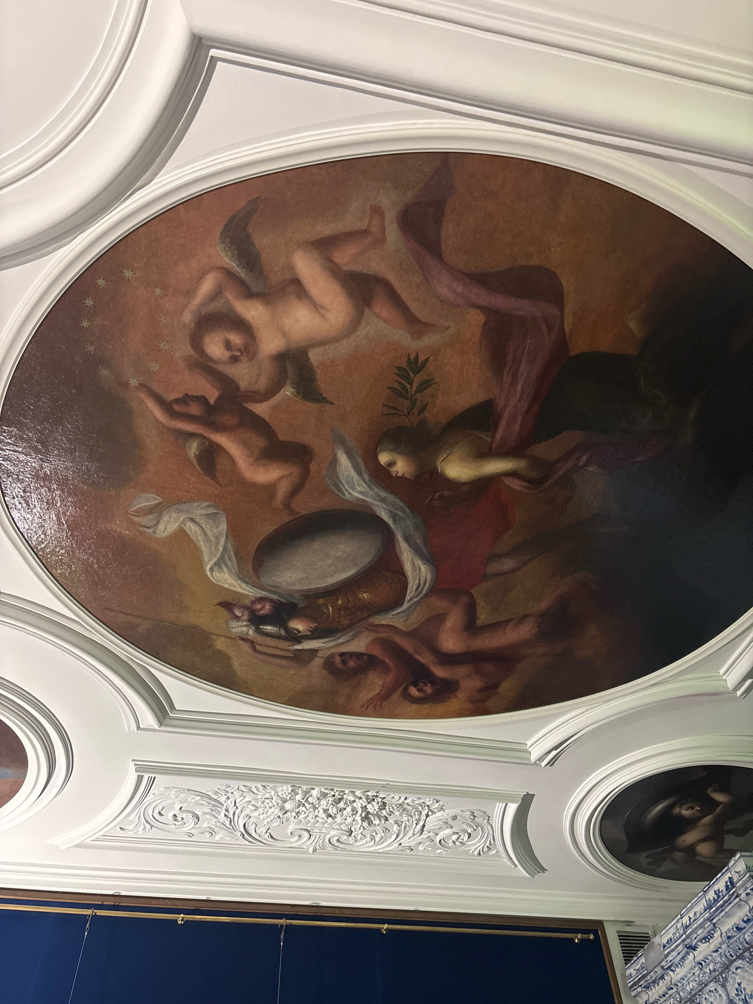 This is a photograph of an ornate ceiling in Saint Petersburg, Russia, featuring a large oval fresco painting surrounded by elaborate white stucco moldings. The fresco depicts several cherubic figures and at least one female figure, rendered in a classical, allegorical style. The figures appear to be in motion, with flowing drapery and dynamic poses, set against a warm, reddish-brown background with scattered star-like elements in the upper left quadrant. In the lower portion of the fresco, a large, dark, concave object, possibly a shield or a drum, is prominently featured. To the right and slightly below the main oval, a smaller, circular fresco depicts a single figure in a darker, more dramatic setting. The surrounding stucco work is intricate, with floral motifs and curved architectural details. Below the ceiling, a dark blue wall is visible, along with a wooden beam and some hanging wires. The overall impression is one of opulence and artistic grandeur, typical of imperial palaces or grand residences in the city.