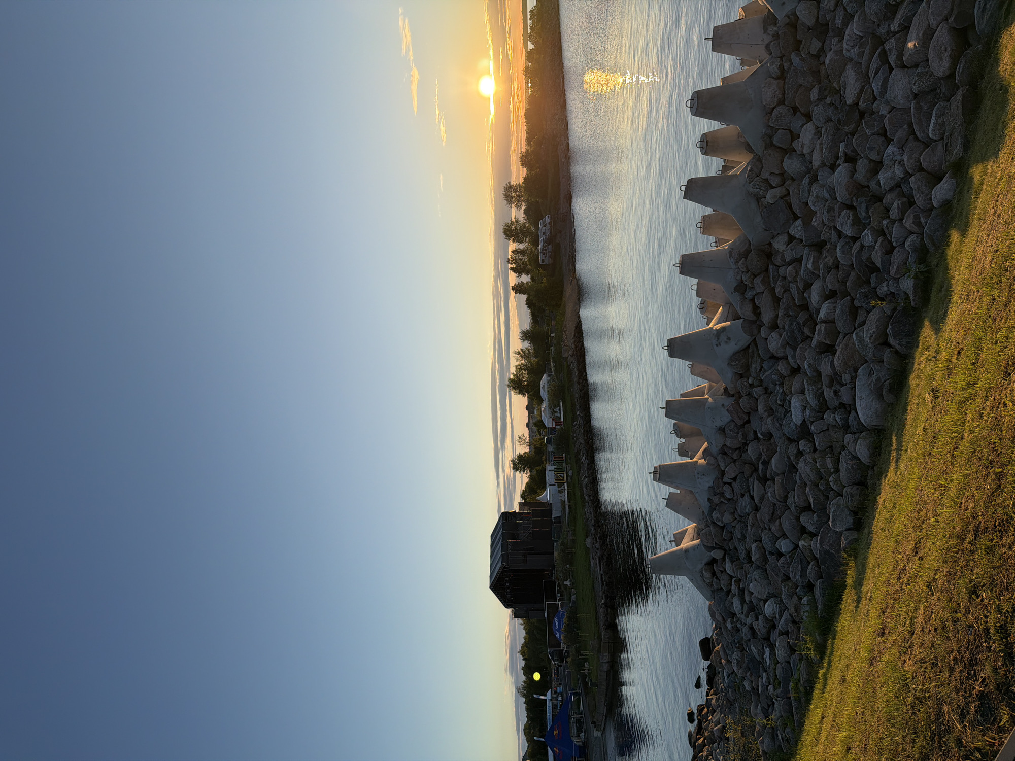 The image depicts a sunset over the water at a location in Tallinn, Estonia. In the foreground, a rocky shore is lined with a series of concrete tetrapods, used as breakwaters. Lush green grass covers the embankment to the left. The water is calm, reflecting the golden hues of the setting sun. Across the water, a dark, large stage structure is visible on the opposite shore, suggesting it might be an outdoor event venue or a park. Several tents and smaller structures are also present in the background, along with trees. The sky is a clear blue, transitioning to warmer tones near the horizon where the sun is setting. No people are visible in the scene.