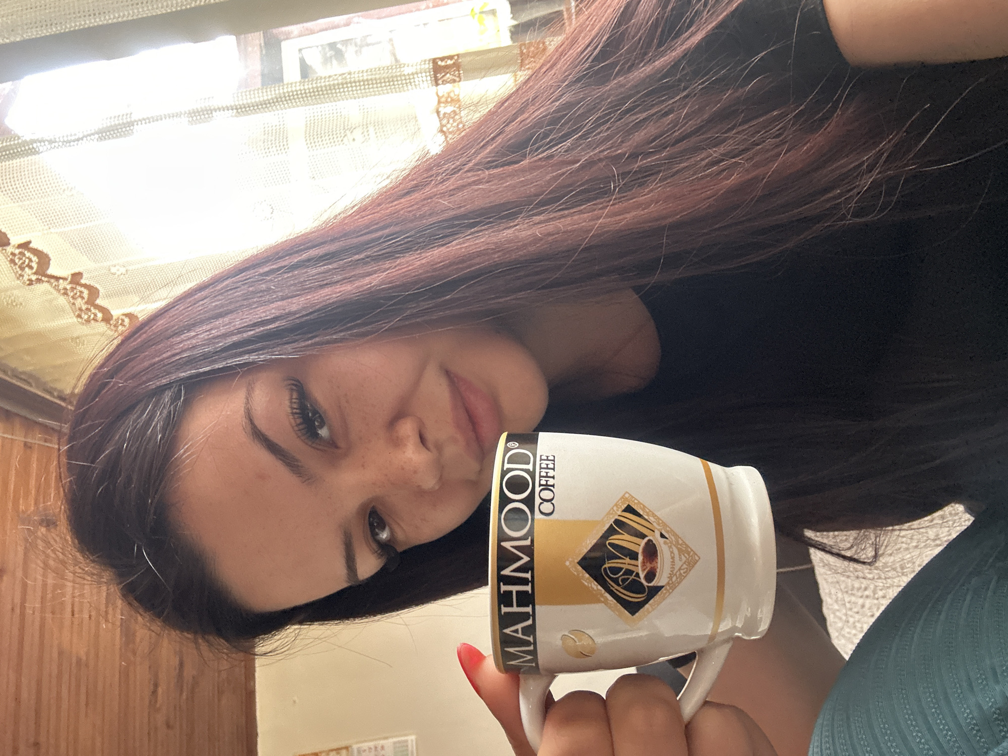 A young woman with long, dark auburn hair and freckles is holding a white ceramic coffee mug with the brand name "MAHMOOD" printed on it. The woman is looking down at the camera with a slight smile. She is wearing a black shirt. Her fingernails are painted red. The background shows a window with lace curtains and sunlight streaming through. A portion of a blue-green fabric is visible in the lower right corner. A wooden surface and a piece of paper are also visible in the background. The location is Chubra, Bulgaria.