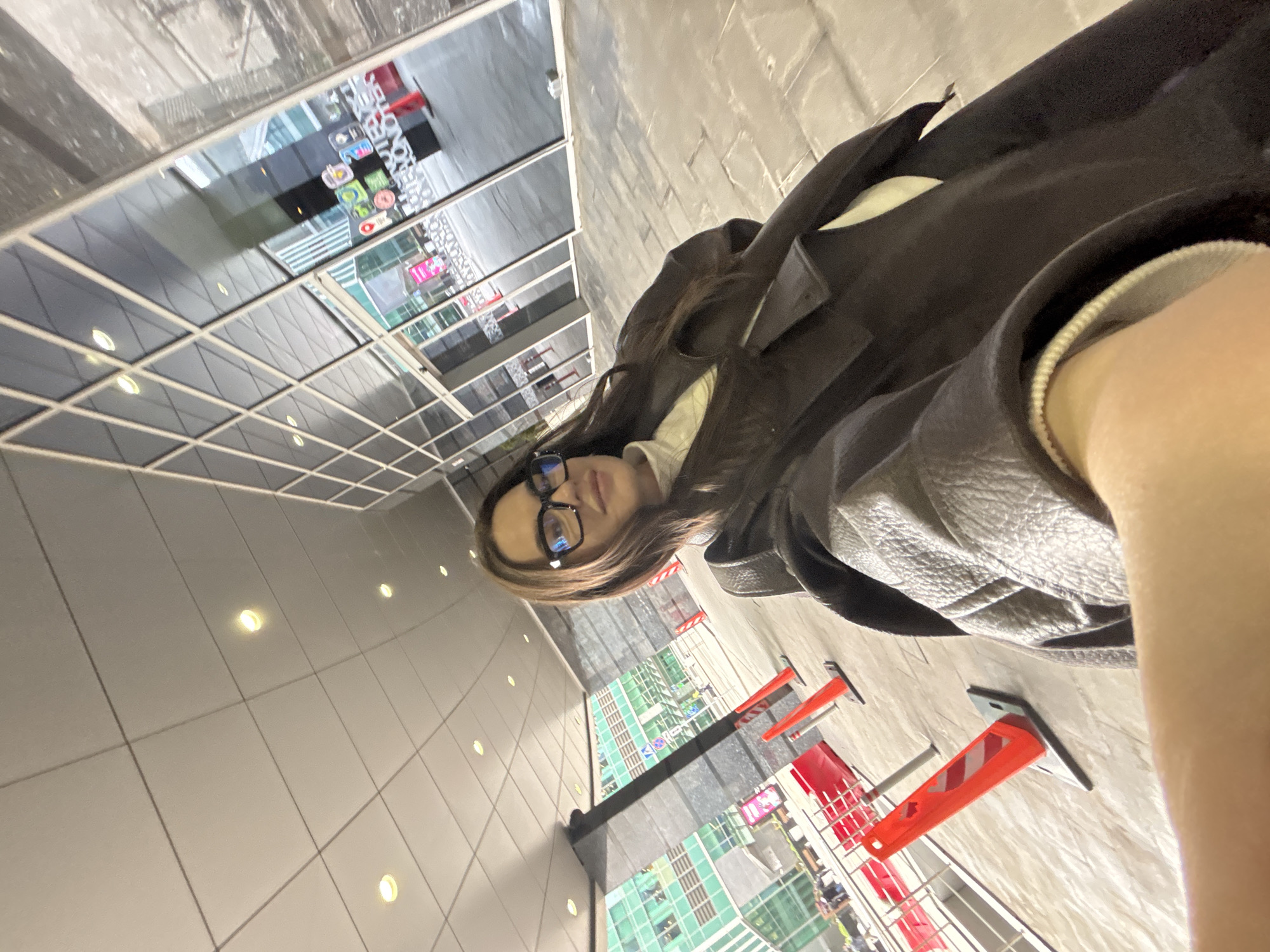 A woman wearing glasses and a dark jacket stands in an indoor space with light-colored tile walls and floors. The scene is likely in Moscow, Russia, given reflections of buildings with Cyrillic signage visible in the windows. Orange and white construction cones are present. The woman appears to be taking a selfie.