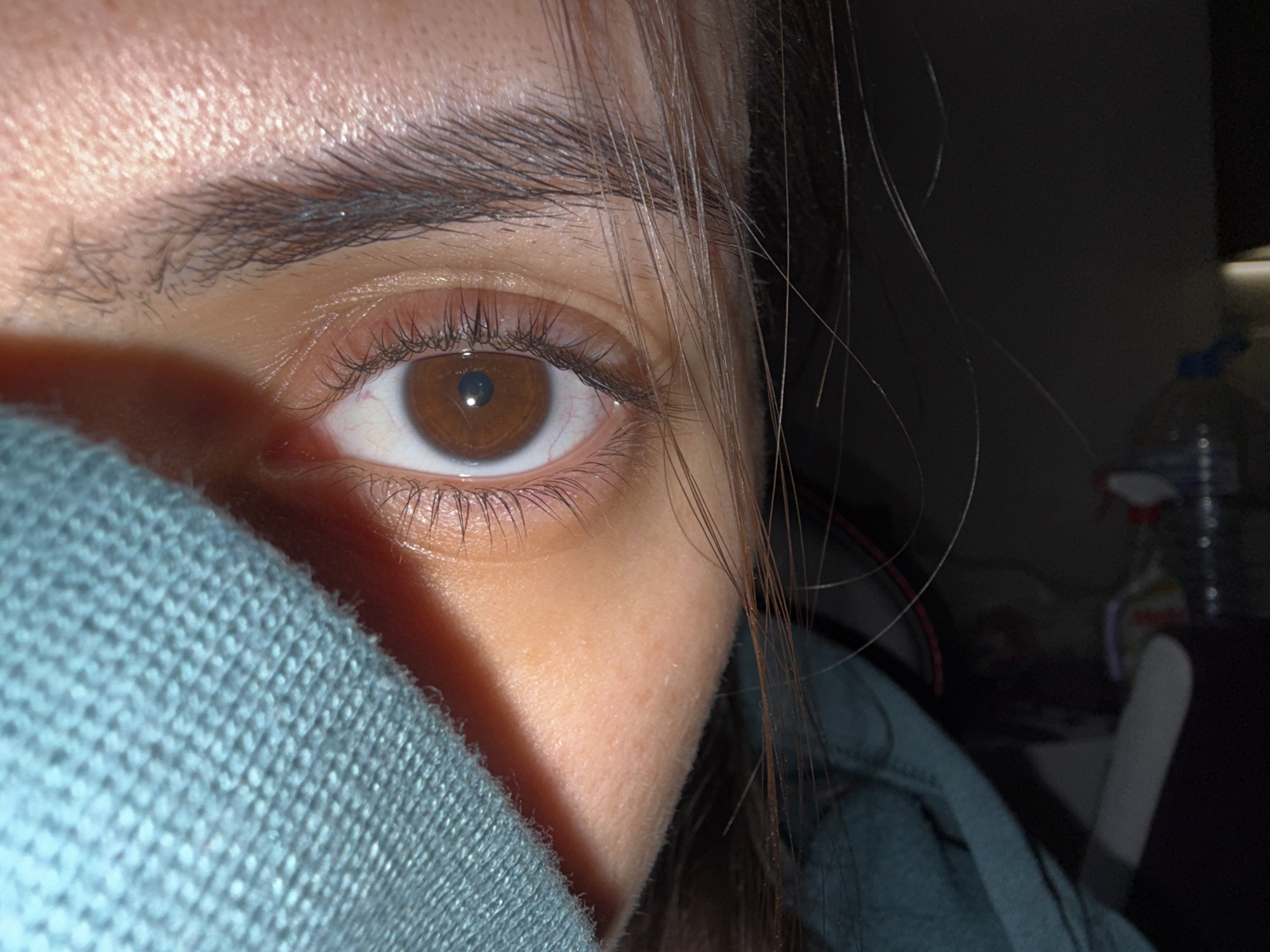 The image shows a close-up of a person's left eye and eyebrow. The eye is brown, and the skin around the eye appears light-skinned. Part of a blue, knitted garment is covering the lower left portion of the face. Strands of brown hair are visible near the eye. The background is dark and indistinct, although a bottle and other objects can be partially seen. There is no information to indicate that the image was taken in Plovdiv, Bulgaria.