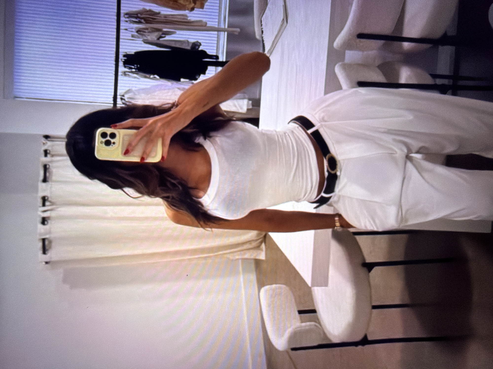 This image shows a woman taking a mirror selfie. She is wearing a white tank top, white pants, and a black belt with a gold buckle. She is holding a light yellow smartphone in her right hand. The background appears to be a well-lit room with white furniture and clothing racks visible. There are also white chairs in the foreground. The woman has long, dark hair and red nail polish. The lighting suggests it could be daytime, and there are no indications of specific weather conditions. The setting seems to be a dressing room or a boutique. There is no visible text in the image.