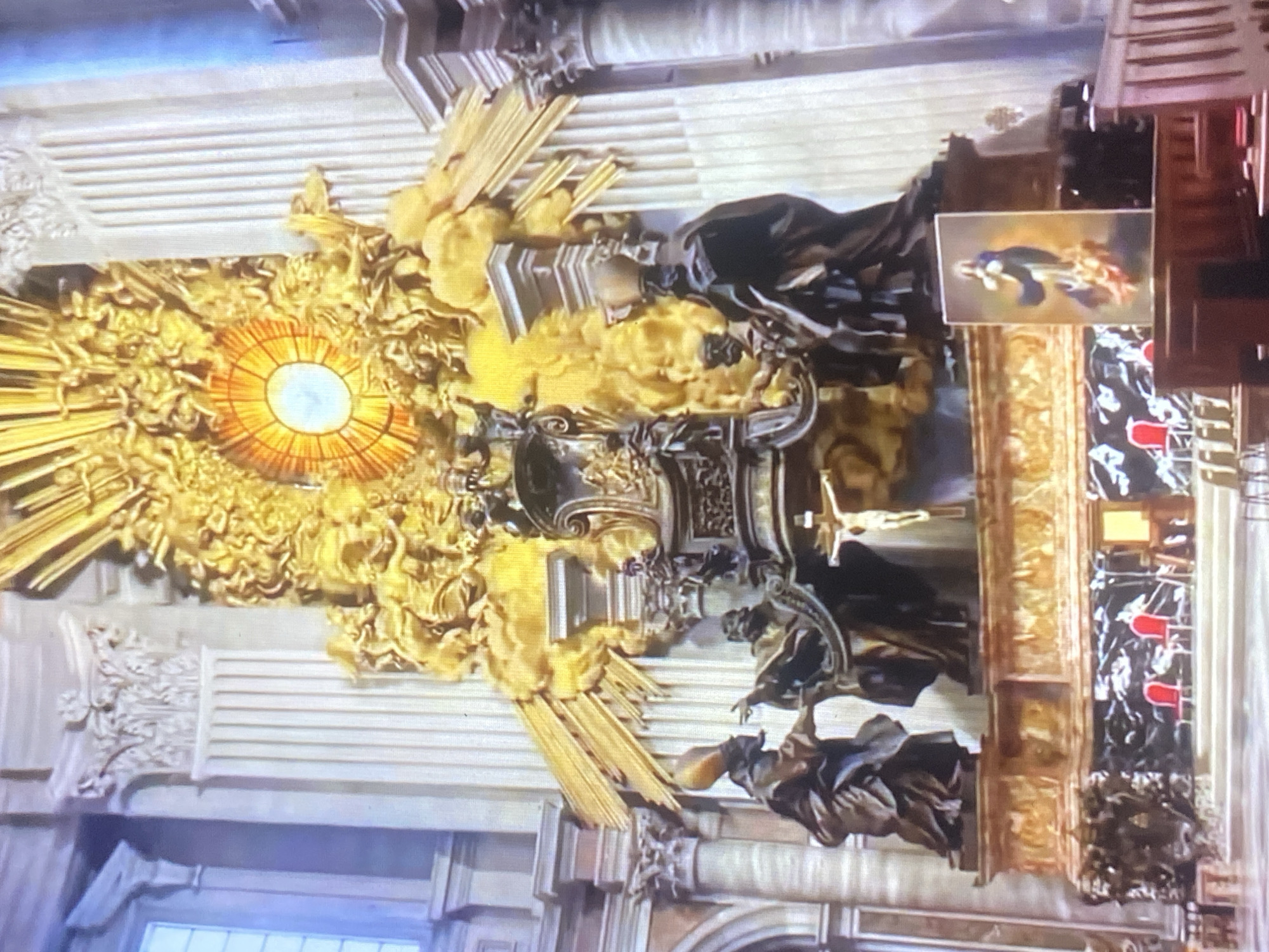 The image displays a richly ornamented interior, likely part of a church or cathedral. The primary focus is a complex, golden sculpture featuring a central radiating light (a "glory"). Statues of figures in flowing robes are placed to the sides, while architectural elements like columns, arches, and decorative panels can be seen in the background. There is a painting in the top right corner of the image. The image is rotated clockwise. The picture has no association with the city of Tinaquillo, Venezuela.