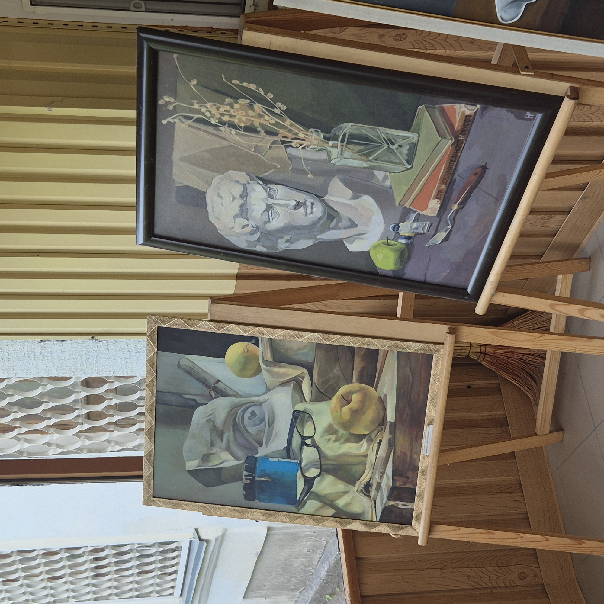Two framed paintings are displayed on wooden easels outdoors, in front of a yellow, ribbed wall and a window with decorative metalwork. The painting on the left features a still life with a plaster bust, a blue container, a pair of glasses, and two apples. The painting on the right depicts a plaster bust of a male head, a glass vase with dried flowers, and a stack of books. The overall scene is an outdoor display of artwork, possibly at a market or exhibition. There are no people visible in the image. The location is Kokshetau, Kazakhstan.