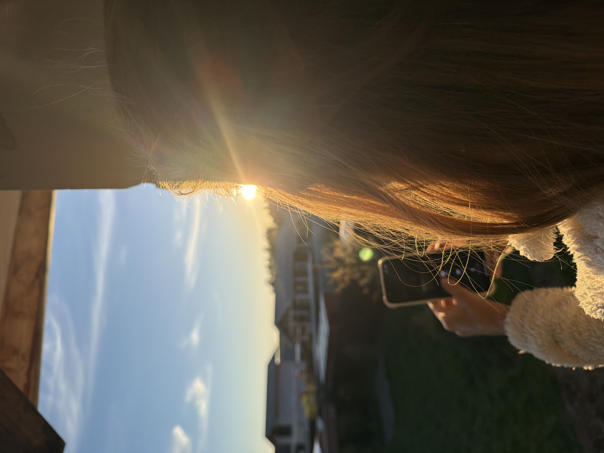 The image shows a person outdoors, holding a smartphone and taking a picture of the sunset. The sun is positioned low in the sky, shining through the person's hair, which is partially obscuring the view. A building is visible in the background along with some trees and what could be utility poles. The sky is blue with some clouds and yellow to the horizon line. The person is wearing a white, fluffy jacket. The location is Kerro Massiv, Russia.