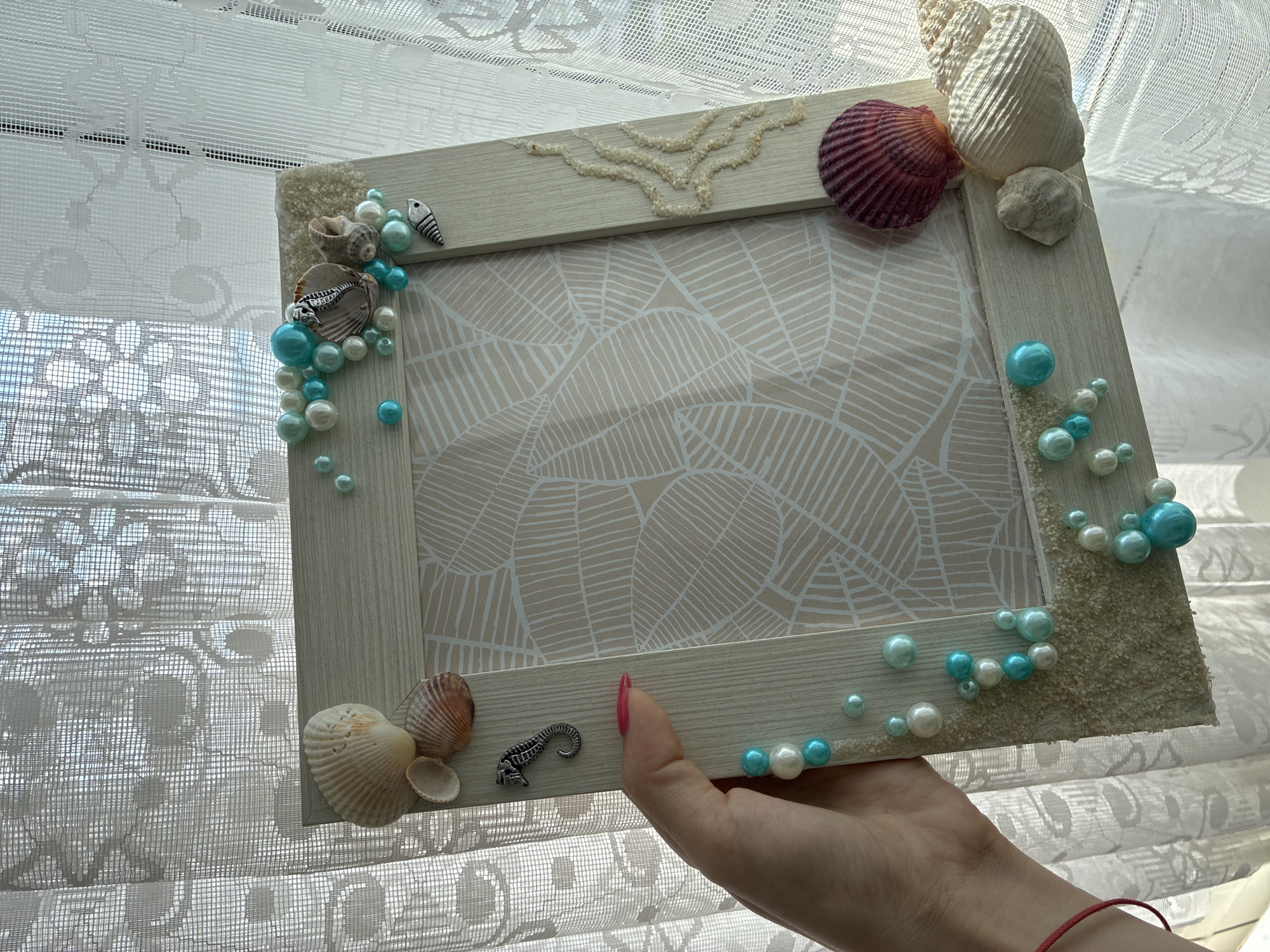 A hand with a pink fingernail is holding a light-colored, wooden picture frame adorned with a nautical theme. The frame is decorated with various seashells, including a large conch shell, a purple scallop shell, and smaller shells. There are also small, shiny pearls or beads in shades of white and teal scattered across the frame, along with small sand-like embellishments and a tiny silver seahorse charm. A small, decorative fish charm is also visible. The background of the frame features a pattern of beige leaves on a white background. Behind the frame, sheer white curtains are visible, allowing sunlight to stream through. The image appears to be taken indoors, near a window. There is no information to identify the location in Burgas, Bulgaria.