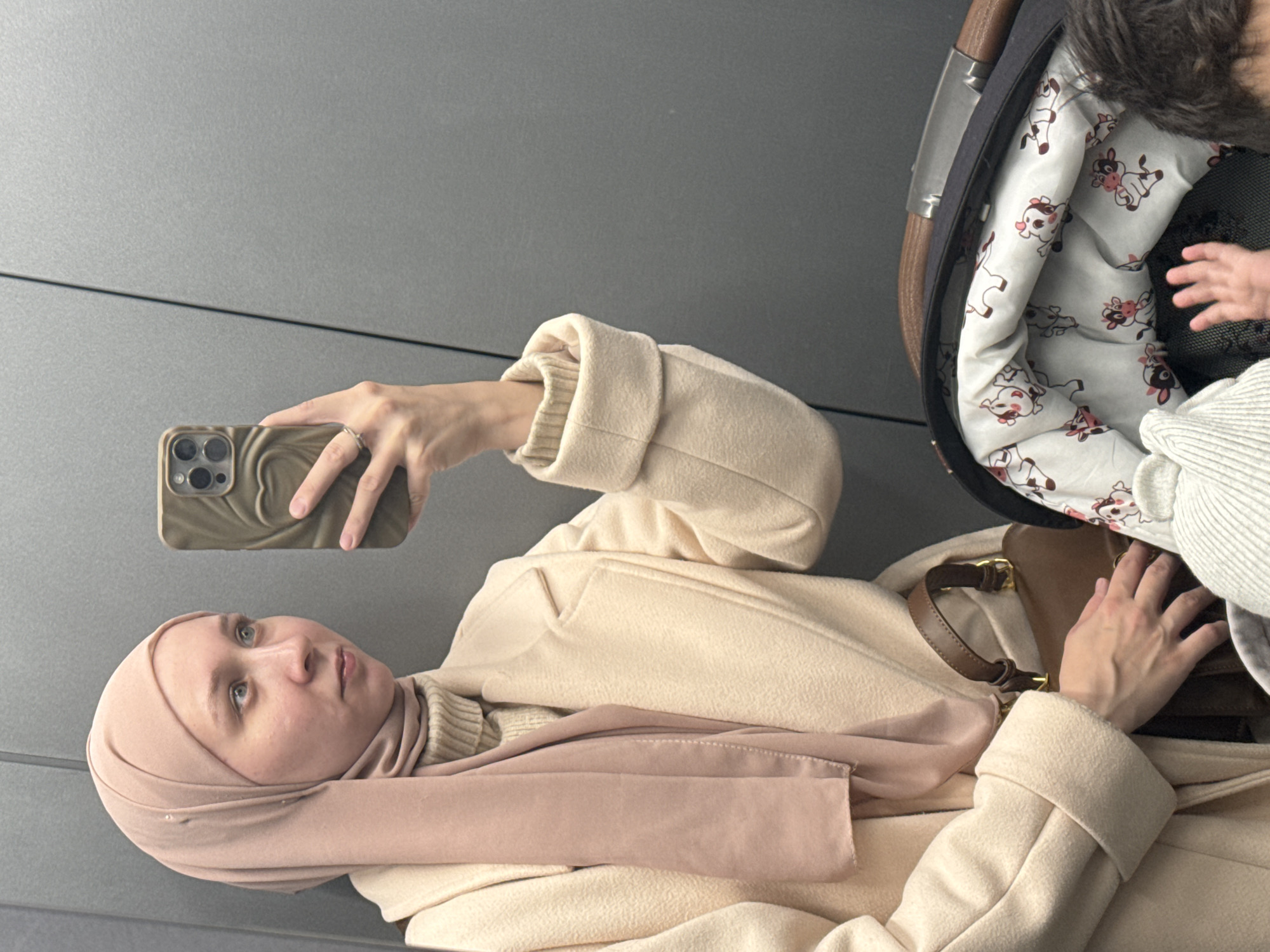 A woman wearing a beige coat and a pink hijab is taking a selfie with her phone. To her right is a baby in a stroller. The background appears to be a gray wall. The location is the city of Chaikovsky, Russia.