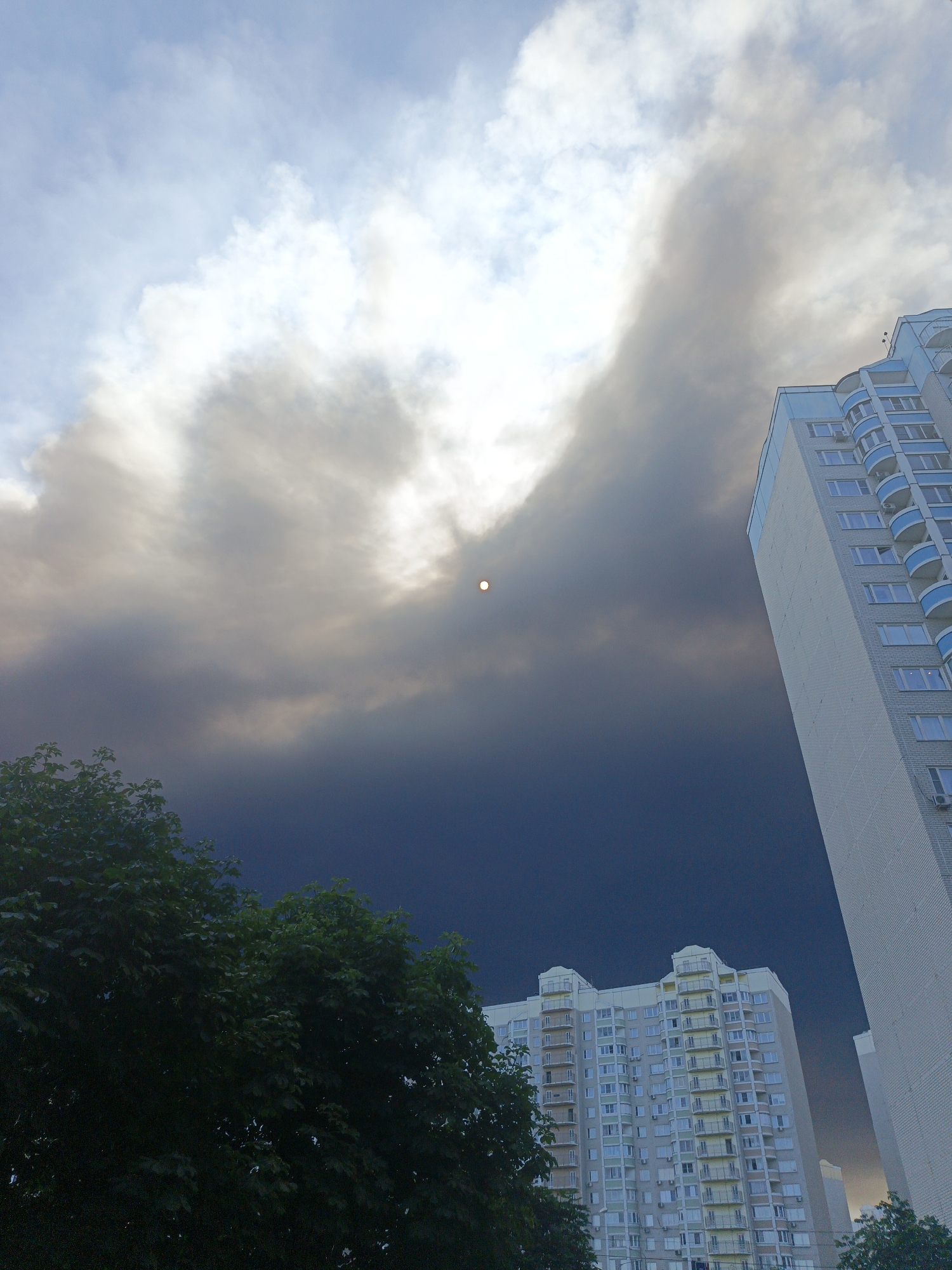 A low-angle shot captures a dramatic sky over Vidnoye, Russia, dominated by dark, swirling clouds that obscure the sun, leaving only a small, bright orb visible. Two tall, modern apartment buildings flank the scene. The building on the right, a light-colored, multi-story structure, features numerous windows and balconies. The building on the left is a lighter, more colorful apartment complex with a distinctive roofline and many balconies. In the foreground, lush green trees are visible, adding a natural element to the urban landscape. The overall impression is one of impending weather change, with the city's architecture set against a turbulent sky.