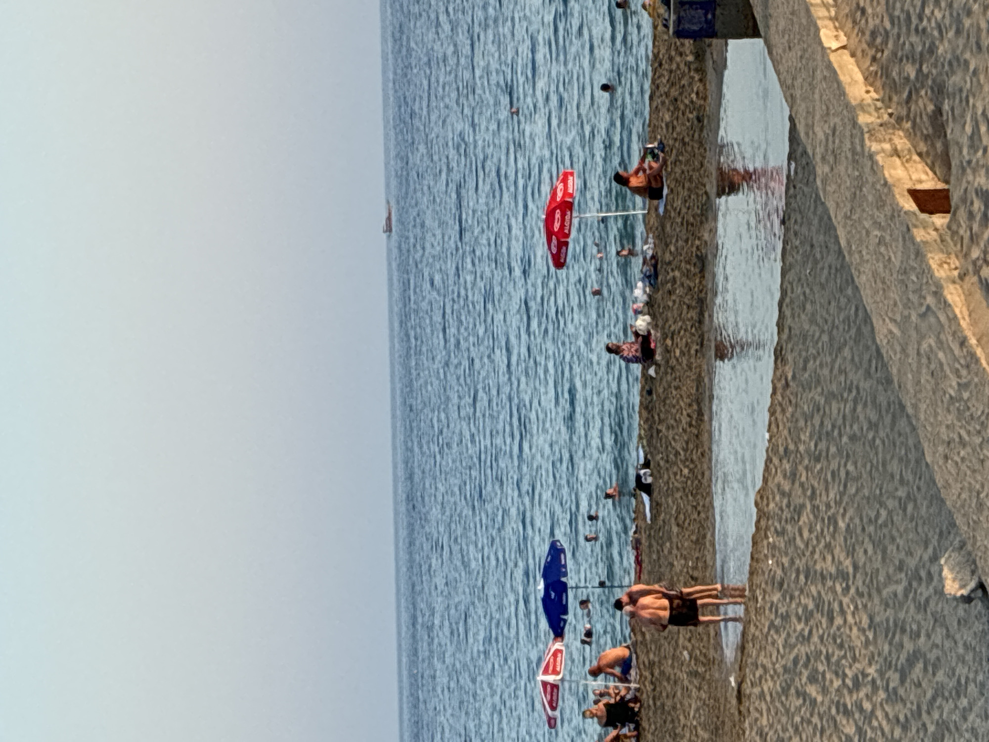 The image captures a serene beach scene in Alanya, Turkey, likely during daylight hours due to the clear sky and soft lighting. The foreground features a sandy beach with some scattered debris. A small, shallow pool of water is visible on the sand, reflecting the sky and surrounding objects.
Further back, several beach umbrellas provide shade to people lounging and swimming. Two red and white striped umbrellas and one blue umbrella are prominent. Numerous individuals are depicted in and around the water, engaged in swimming and sunbathing. Some are partially submerged in the sea, while others are seated on the sand.
In the mid-ground, a few people are visible near the umbrellas, some appearing to be in conversation or relaxing. One individual is sitting under a red umbrella, appearing to be packing up belongings. Further out in the water, other swimmers can be seen.
The sea is a prominent feature, with gentle waves and a clear horizon. A small boat or vessel is visible in the distance on the water. The sky is a pale blue, transitioning to a lighter shade towards the top of the frame. The overall atmosphere is one of leisure and relaxation on a typical beach day.
