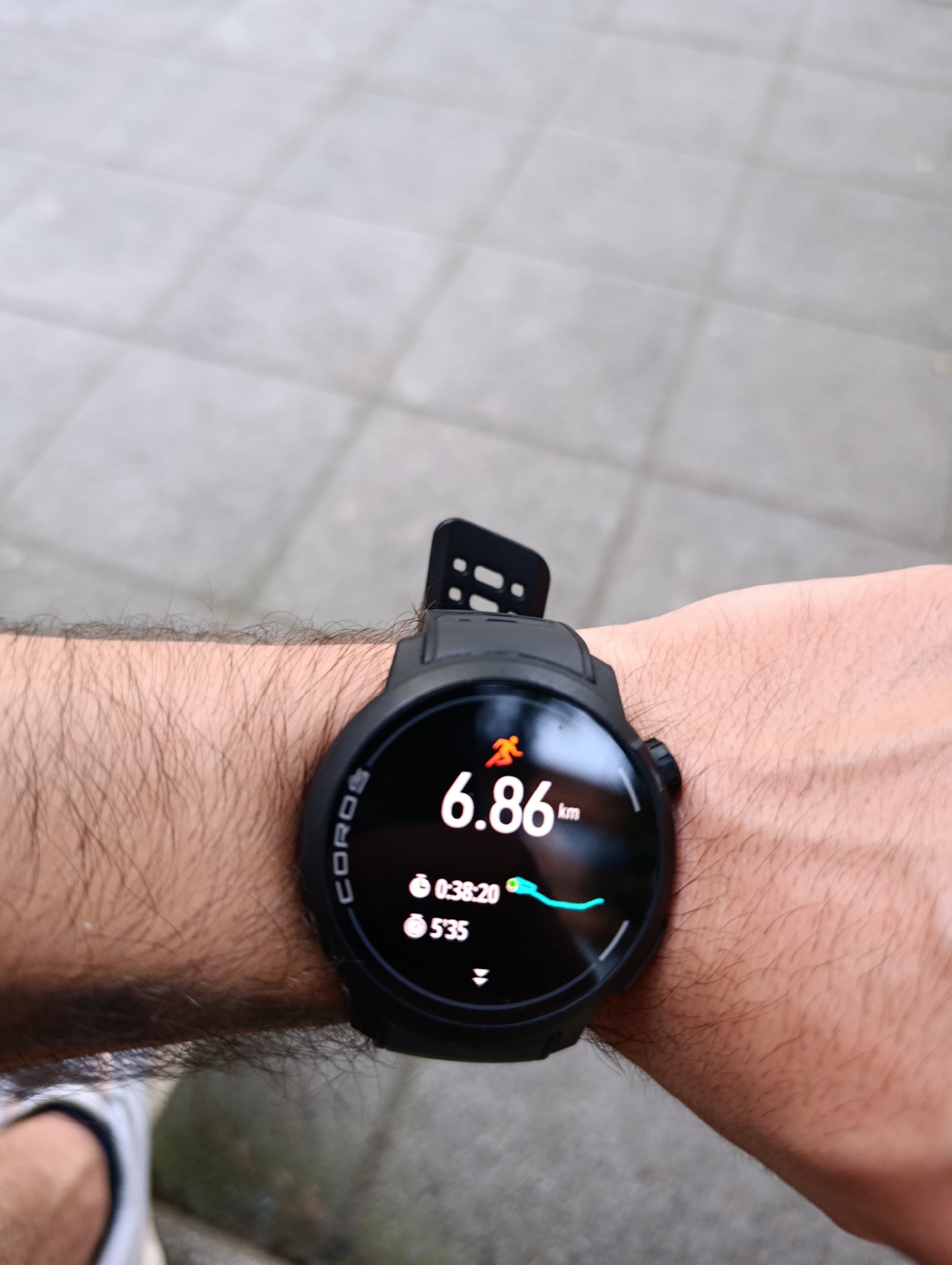 A person's hairy arm is visible, wearing a black Coros smartwatch on their wrist. The watch face displays an activity tracking screen. Prominently shown are the distance "6.86 km", a running icon, and elapsed time "0:38:20". Below that, "5'35" is displayed, likely indicating pace. A green line on the screen suggests a mapped route. The background is a light-colored, textured surface, possibly a paved area with square tiles, common in urban environments. The image's focus is on the smartwatch and the arm, suggesting the context of a workout or outdoor activity. There is no visual information within the image to suggest the location is Razgrad, Bulgaria.