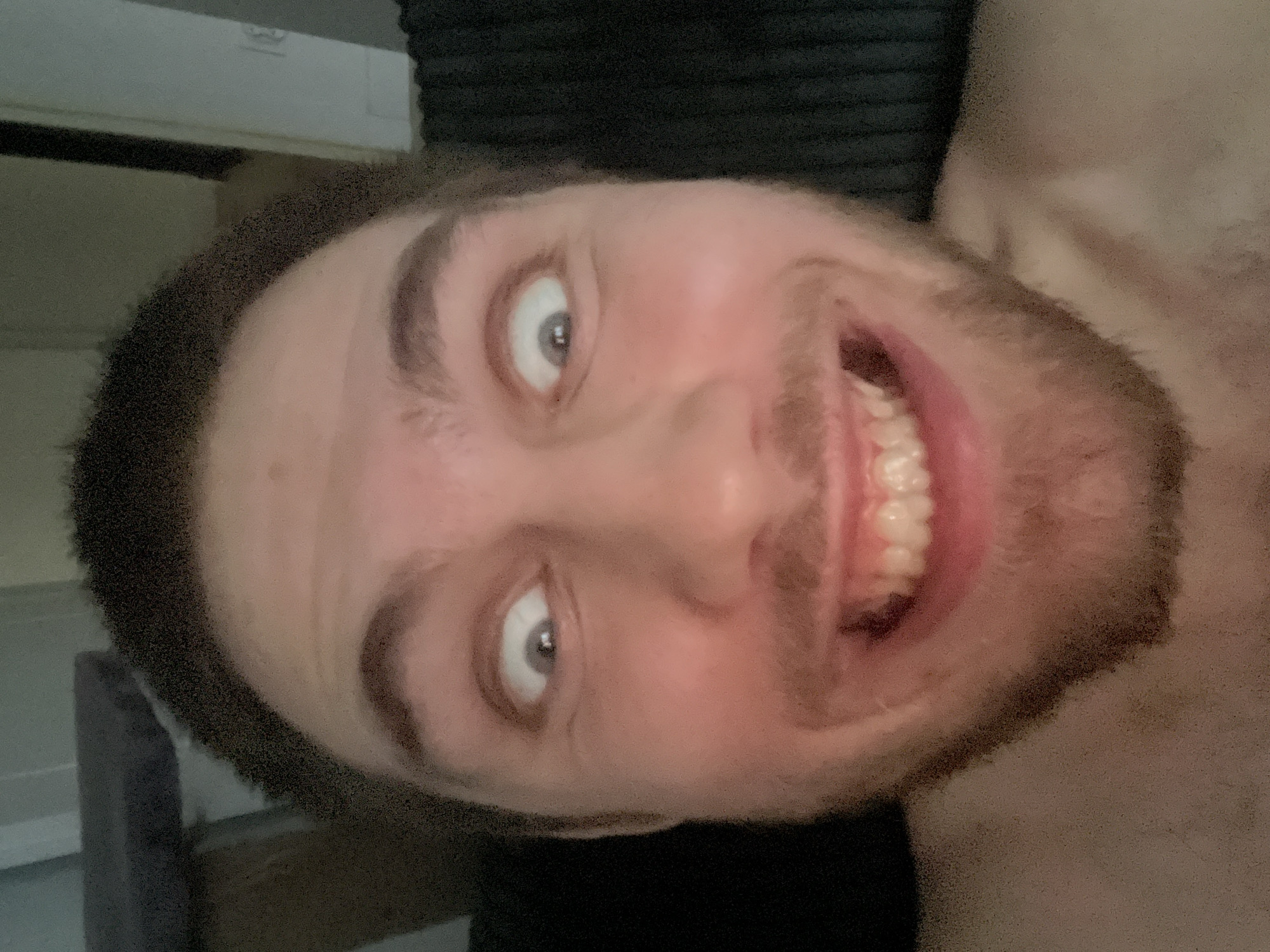 This media file is a selfie, rotated 90 degrees counter-clockwise, of a young man's face. He has light skin, brown hair, and a light beard. He is smiling broadly, showing his teeth, and his eyes are wide open and looking slightly upwards. The image appears to be taken indoors, possibly in a bedroom, given the soft lighting and the presence of what looks like a headboard in the background. The man's expression suggests a playful or jovial mood. There is no visible text in the image. The metadata indicates the location is Pruszcz Gdański, Poland, but this cannot be visually confirmed from the image itself.