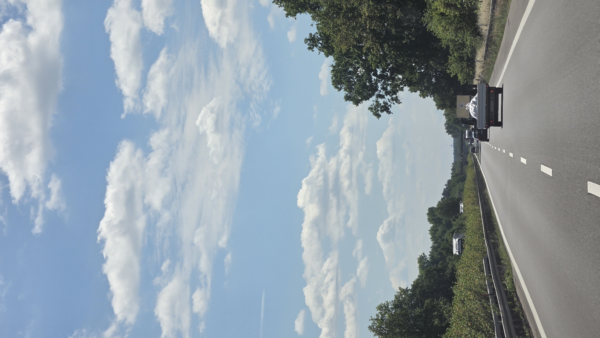This media file shows a view from a vehicle driving on a highway in Germany, likely near Calau. The perspective is from the passenger side, looking forward and slightly upwards. The scene is dominated by a bright blue sky filled with fluffy white clouds. Lush green trees line the right side of the highway, creating a natural barrier. The highway itself is asphalt with white dashed lines marking the lanes.
Several vehicles are visible on the road ahead, suggesting moderate traffic. The closest vehicle is a dark-colored pickup truck with a white tarp covering its cargo bed. Behind it, other cars and trucks are in motion. The time of day appears to be daytime, with bright sunlight casting shadows and illuminating the scene. The weather seems pleasant, with clear skies and scattered clouds.
The overall impression is a typical highway drive through a scenic, green landscape. There is no visible text in the image. The composition emphasizes the open road and the expansive sky, conveying a sense of travel and freedom. The presence of trees suggests a rural or suburban setting.
