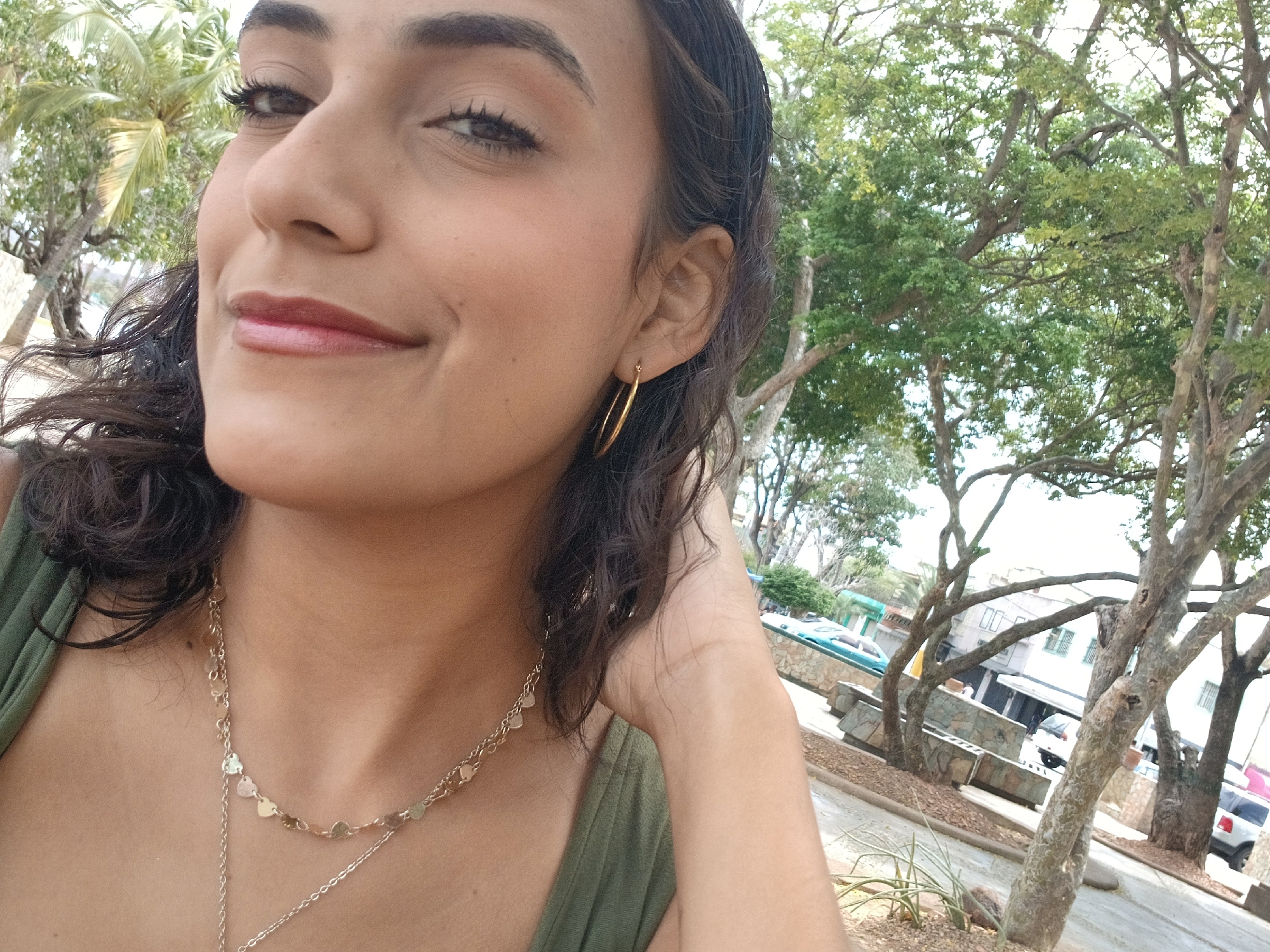 A bright, daytime portrait-style photograph features a young woman smiling outdoors in Juan Griego, Venezuela. The woman, positioned on the left side of the frame and seen from the chest up, gazes upwards and to the viewer's left with a pleasant, relaxed smile. She has dark, curly hair, brown eyes, and appears to be wearing light makeup, including mascara and a glossy, reddish-pink lip color. Her attire includes a green top, a golden hoop earring on her visible right ear, and two layered gold necklaces, one embellished with small heart-shaped charms.
The background reveals a sunny, outdoor setting. Lush green trees, some with broad leaves and others showing more distinct branches, dominate the upper and right portions of the image, suggesting a tropical or subtropical environment consistent with Venezuela. In the mid-ground and distance, elements of an urban or town area are visible through the foliage, including buildings, a paved street or pathway, and low stone walls or planters. A white car is partially visible in the far right background. The clear lighting indicates it is daytime. No text is visible in the image.