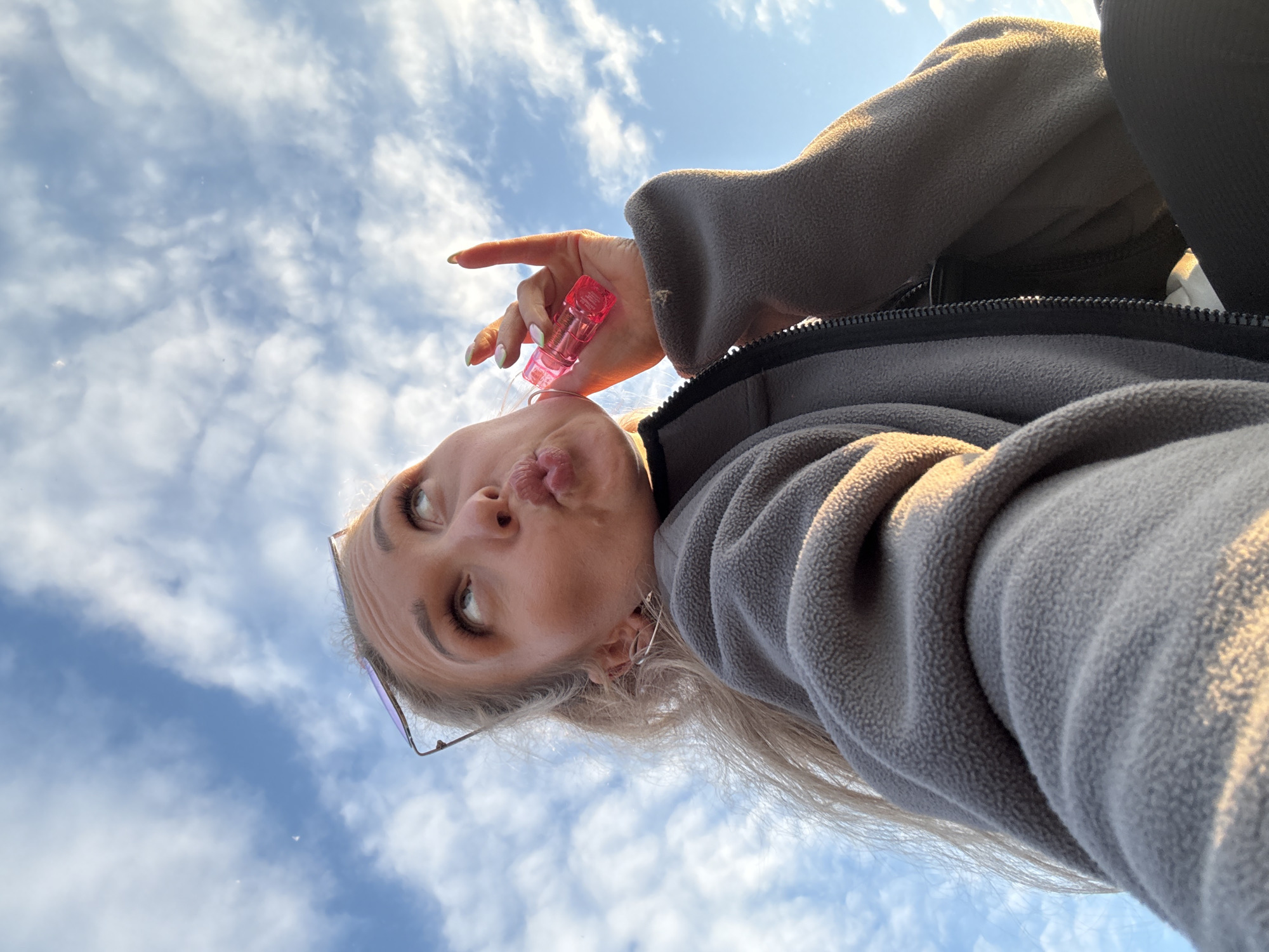 A low-angle selfie captures a young adult female positioned against a clear blue sky sparsely populated with white cumulus clouds, illuminated by warm, directional light. The subject's face is turned upwards and slightly to the viewer's left, with her eyes directed similarly. Her lips are puckered in a "kiss face" expression. Sunglasses are perched on her blonde hair, which is styled in a ponytail, with a few strands framing her face. She wears thin, silver-colored hoop earrings. Her right hand (viewer's left) is raised, revealing long, almond-shaped fingernails painted with light-colored polish, holding a small, translucent pink cylindrical object. She is dressed in a grey fleece jacket featuring a visible black zipper and a dark-colored garment or strap beneath. The specific city of Chaikovsky, Russia, cannot be determined from the visual information presented in the image.