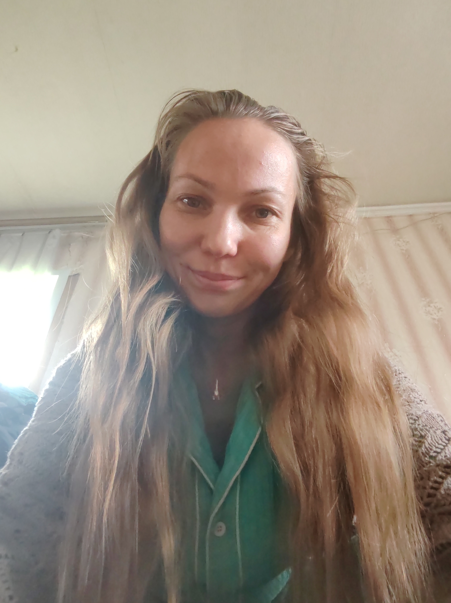 A woman with long, naturally wavy, light brown hair is depicted in a close-up, likely a self-portrait, looking directly at the camera with a gentle, calm smile. Her hair flows over her shoulders and chest. She has fair skin and brown eyes.
She is wearing a teal or light green collared top with white piping, and a delicate pendant necklace is visible around her neck. A light-colored, possibly beige, knitted or crocheted garment is draped over her left shoulder.
The setting is indoors, with a bright natural light source, likely a window, illuminating the left side of the frame, suggesting it is daytime. The background consists of a light-colored wall; a section on the right shows subtle patterned wallpaper. The ceiling is visible at the top. No text is visible in the image. The scene conveys a relaxed, informal atmosphere within a residential interior in Чухлэм, Russia.