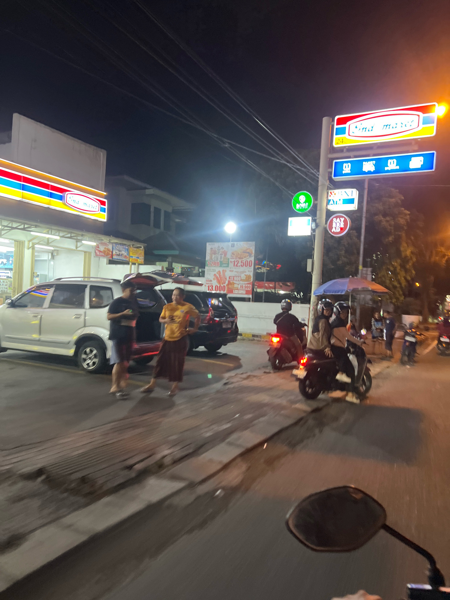 At night, in front of an Indomaret convenience store in Magelang, Indonesia, several people are gathered. A silver minivan with its trunk open is parked on the left, and a dark-colored SUV is behind it. Two men, one wearing a dark t-shirt and shorts, and another in a yellow t-shirt and a long skirt or sarong, stand by the minivan.
Several motorcycles are present. A motorcycle with two riders, both wearing helmets, is in the middle of the road. Another motorcycle is parked to the right, and a third motorcycle is visible further down the street. On the right side of the image, a street vendor's cart with an umbrella is set up.
A street sign with the Indomaret logo is illuminated, and below it are signs indicating an ATM and other services. Several posters with text and prices are also visible on the building and a pole. The street is wet, reflecting the lights from the store and signs. The overall scene depicts a typical street view at night in an Indonesian city, with a convenience store, vehicles, and people engaged in various activities.