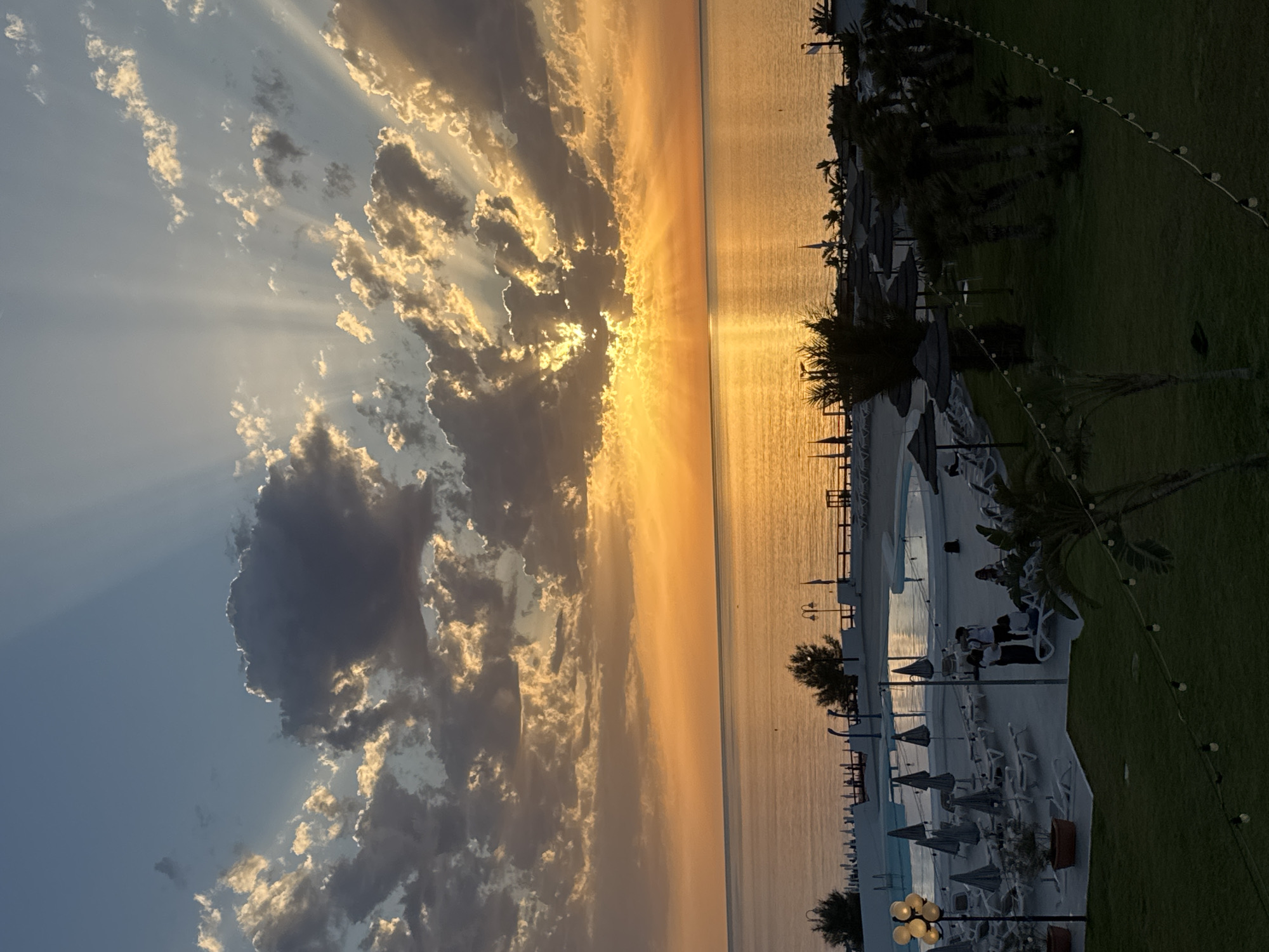 A sunset over the Mediterranean Sea is depicted, with rays of sunlight piercing through dramatic clouds. The scene takes place at a resort or hotel in Isola delle Femmine, Italy. In the foreground, a well-maintained lawn with string lights is visible, leading to a swimming pool area. Numerous lounge chairs and umbrellas are set up around the pool and on the beach. A few people are present, some gathered near the pool and others walking on the deck. Palm trees and other vegetation dot the landscape, adding to the tropical ambiance. The calm sea reflects the vibrant colors of the sunset, creating a serene and picturesque view.