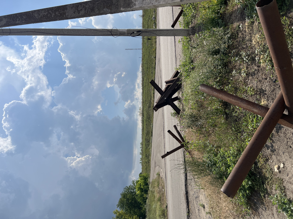 The image depicts a daytime outdoor scene at a road junction in Shestakove, Ukraine, characterized by the presence of defensive anti-tank obstacles. In the immediate foreground, a rusty anti-tank obstacle constructed from three intersecting metal pipes lies on the ground, with two pipes extending towards the viewer. The ground in the foreground consists of dry soil and sparse green vegetation.
In the midground, a paved asphalt road curves from the left towards the center and then to the right, disappearing beyond the frame. To the left of the road, a smaller, upright anti-tank obstacle, composed of three metal bars, is visible amidst green foliage. To the right of the road, a larger, darker anti-tank obstacle, resembling a Czech hedgehog constructed from heavy beams, lies on its side in the grass. Further to the right, two grey concrete utility poles stand vertically, with one pole featuring black electrical cables running along its length. Another single metal pipe, possibly part of a disassembled obstacle, is visible behind the rightmost utility pole.
The background shows a gently rising terrain covered in green vegetation, suggesting an open field or hill. A thin, dark vertical structure, possibly a communication tower, is discernible on the distant horizon line. The sky above is predominantly blue with significant formations of large, bright white and grey cumulus clouds, indicating partial cloud cover. No individuals or active movements are present in the scene.