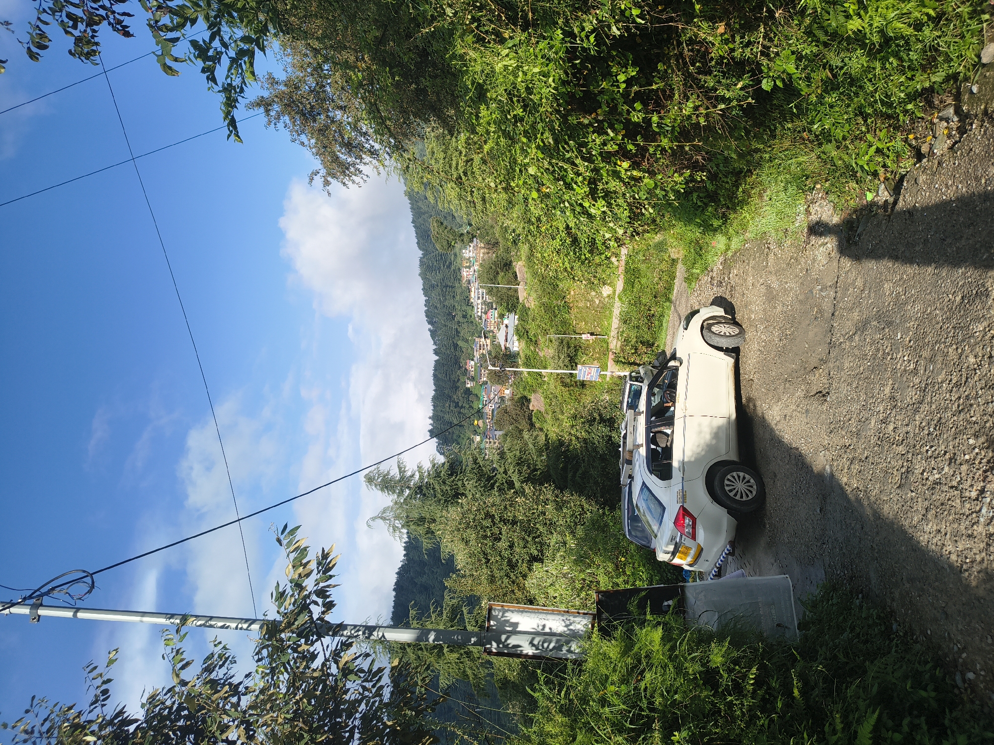 The image depicts a scene in the city of Haini, India, on a sunny day with a clear blue sky and scattered clouds. A rough, gravel road winds through a lush, green landscape. On the left side of the road, a tall, grey utility pole stands with several wires running overhead. Dense green foliage encroaches on the left side of the road, with some leaves showing yellowing.
Parked on the road are two vehicles. The primary subject is a white sedan, likely a taxi, with its rear end facing the viewer. It has a roof rack. A dark-colored car is partially visible behind it.
In the background, a hillside is covered with numerous buildings, suggesting a town or city built on a slope. These buildings are a mix of colors, predominantly white and pastel shades, with red and green roofs. The hillside is densely populated with trees, primarily evergreens, creating a vibrant green backdrop. The overall impression is of a rural or semi-urban setting in a hilly region. A person's shadow is cast on the road in the lower right portion of the frame.